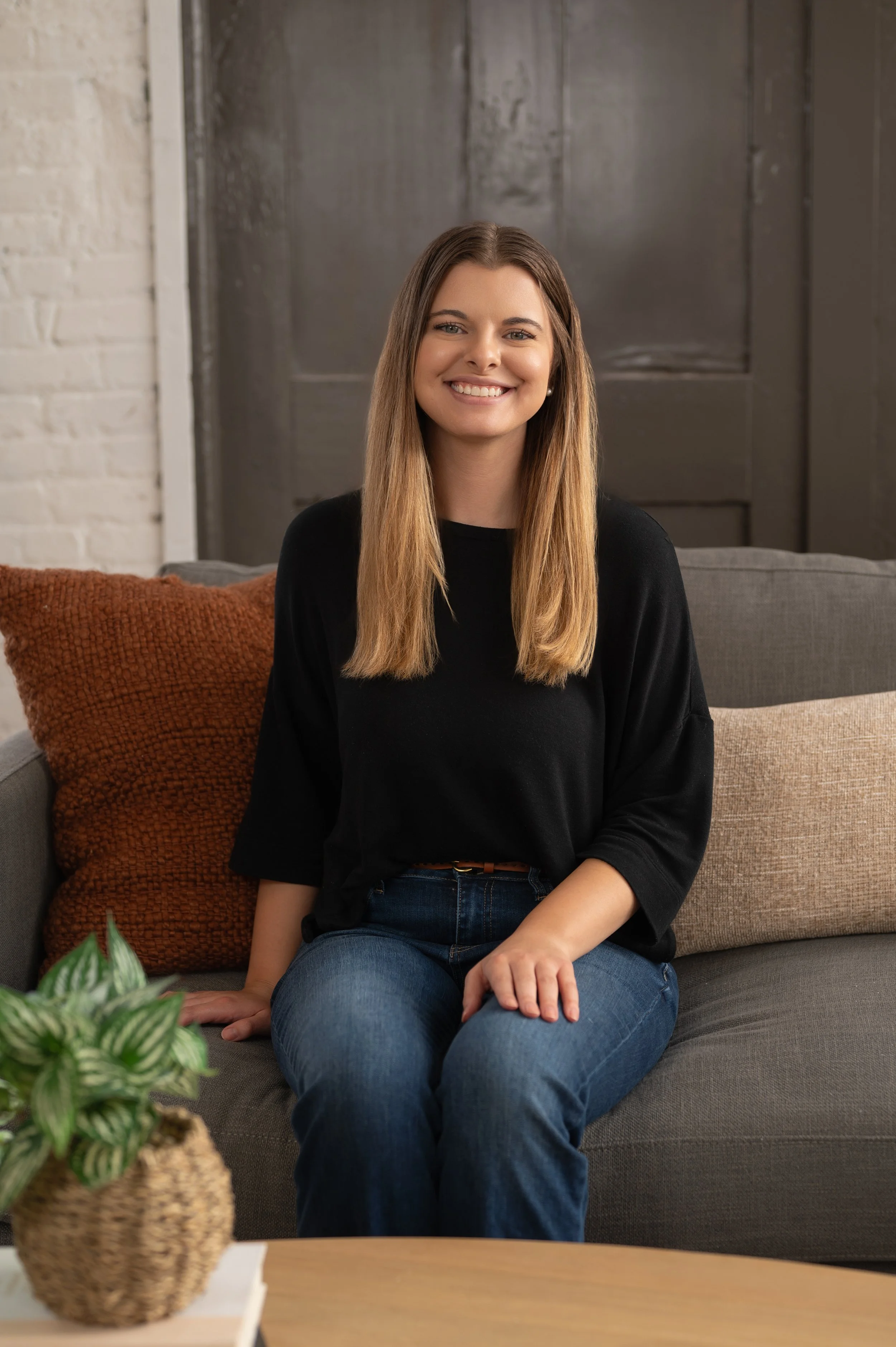 Woman smiling on a gray sofa in a loft with whitewashed brick — a relaxed in-home portrait