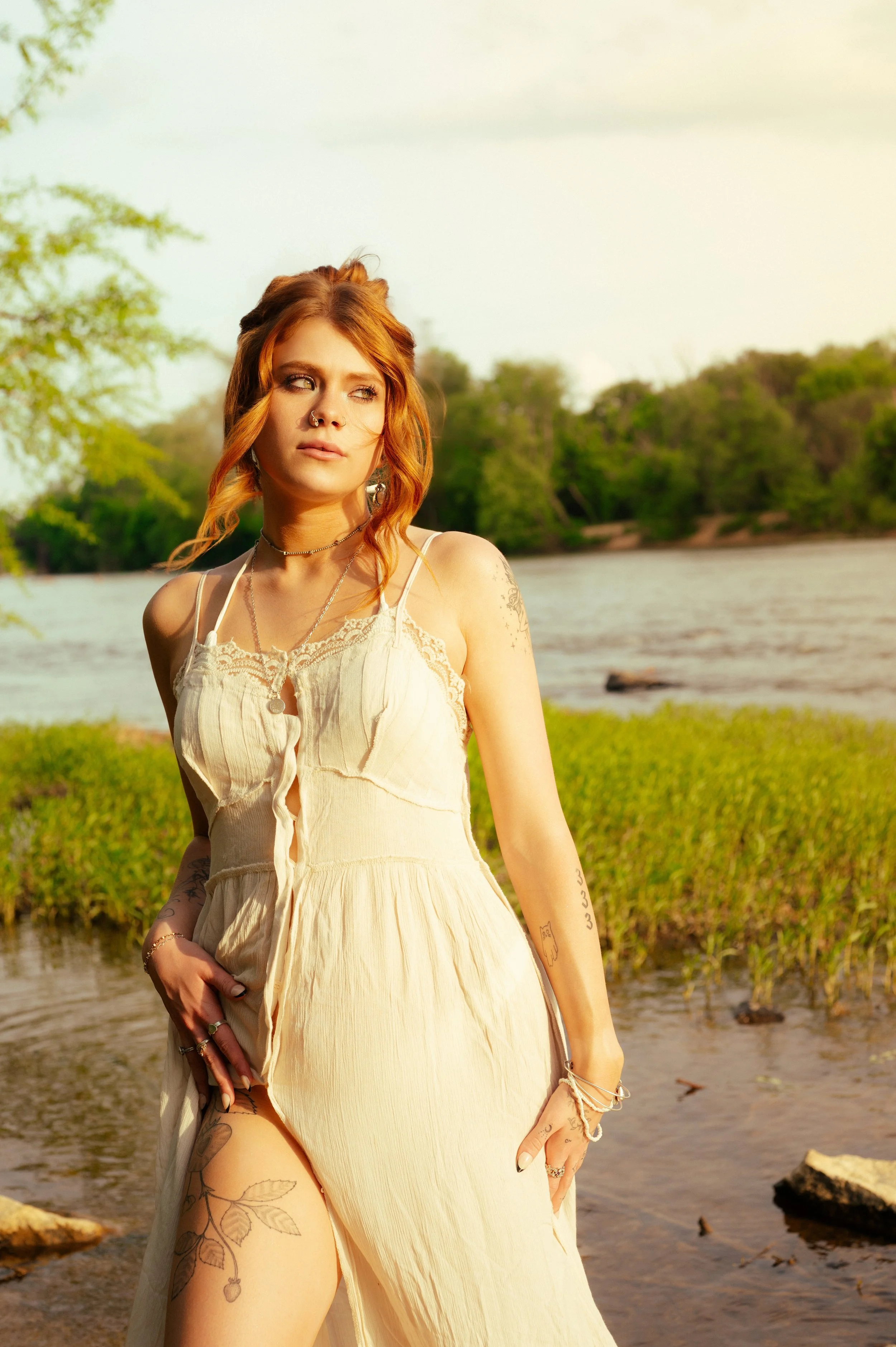 A woman with copper hair in a cream slip dress at the river's edge at golden hour