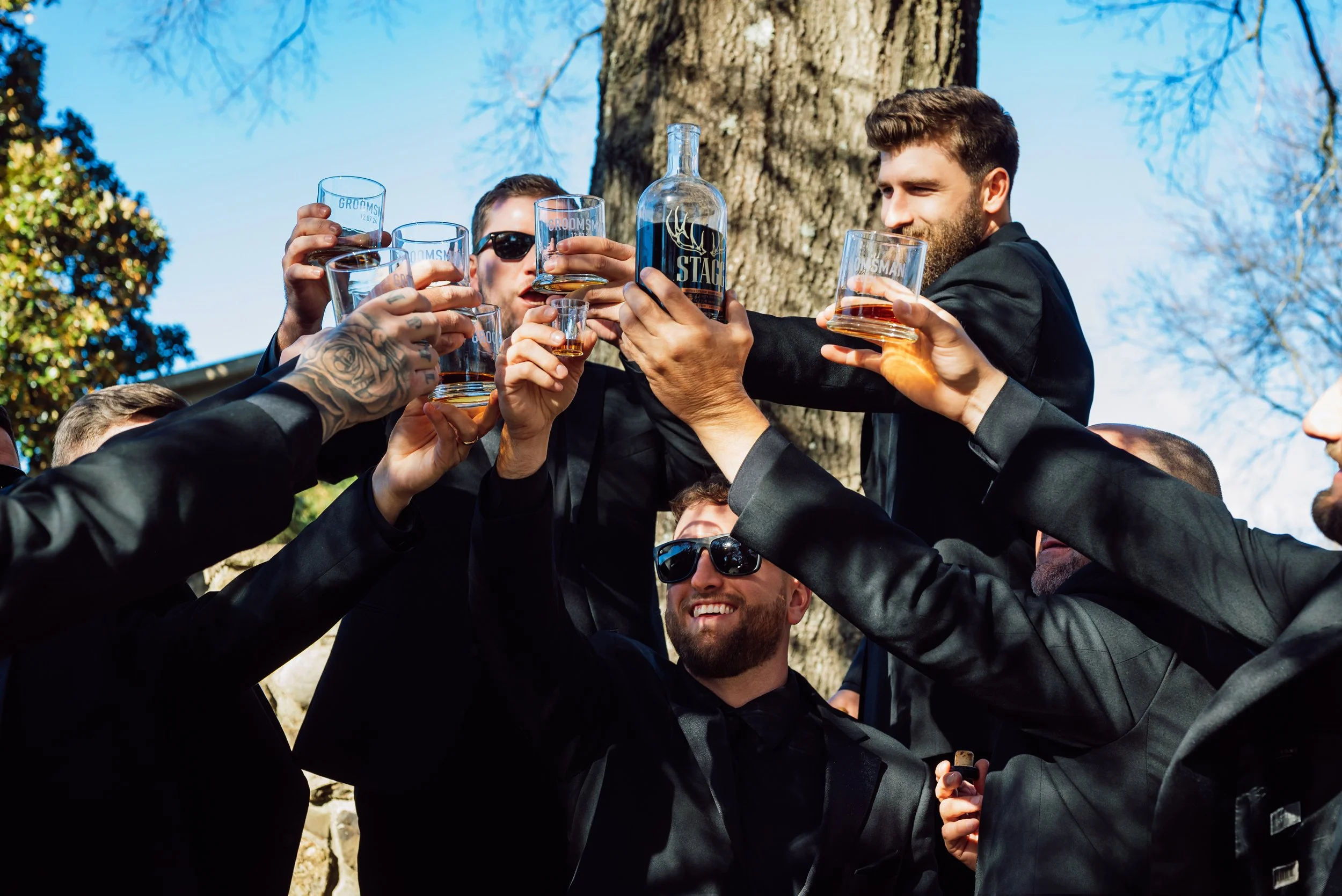 Groomsmen in black suits raising engraved whiskey glasses in a toast beneath a tree at the Adcock wedding