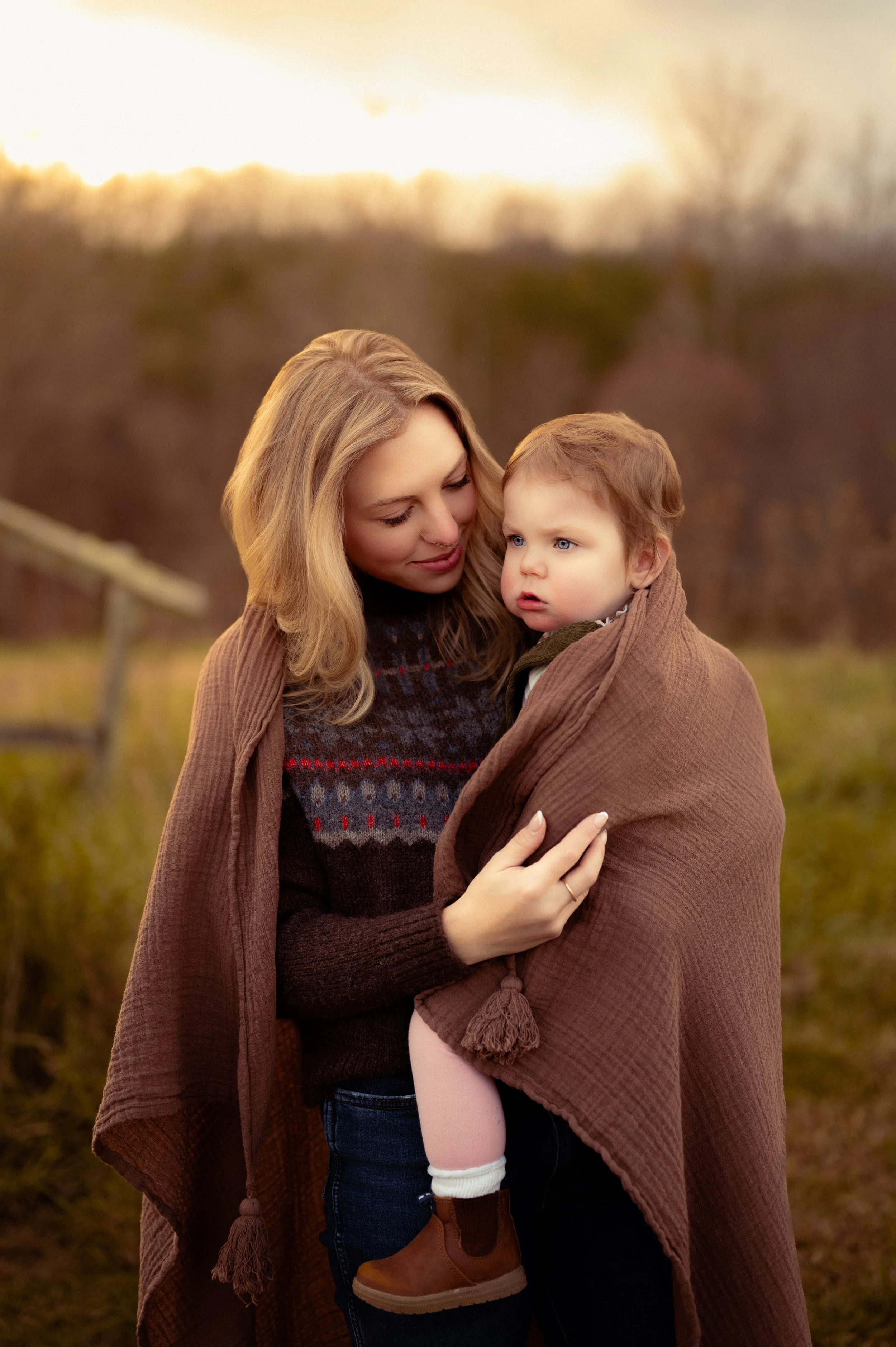 A mother holding her toddler wrapped in a brown blanket in a field at dusk