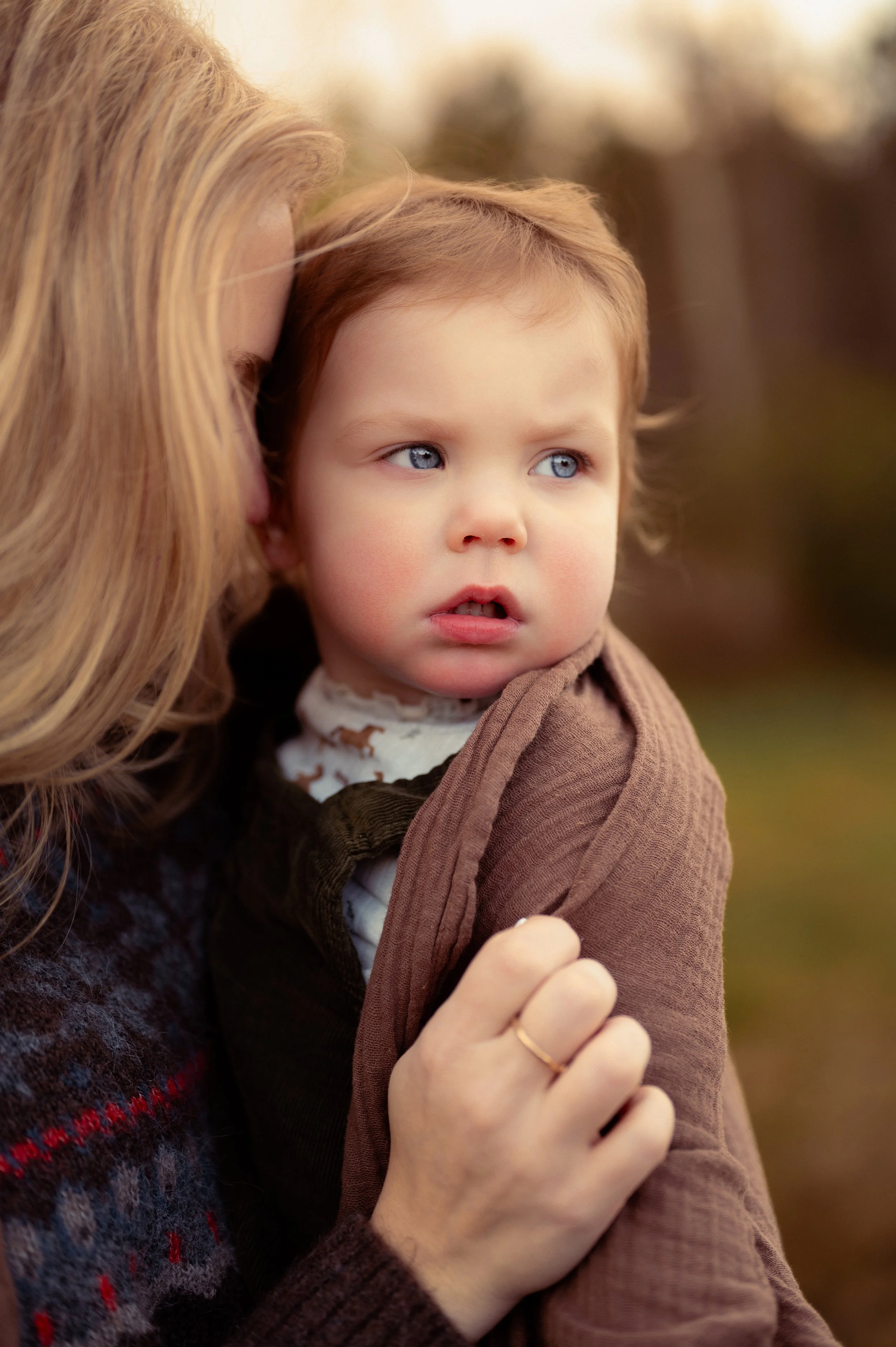 Mother holding her blue-eyed toddler wrapped in a brown muslin blanket at dusk