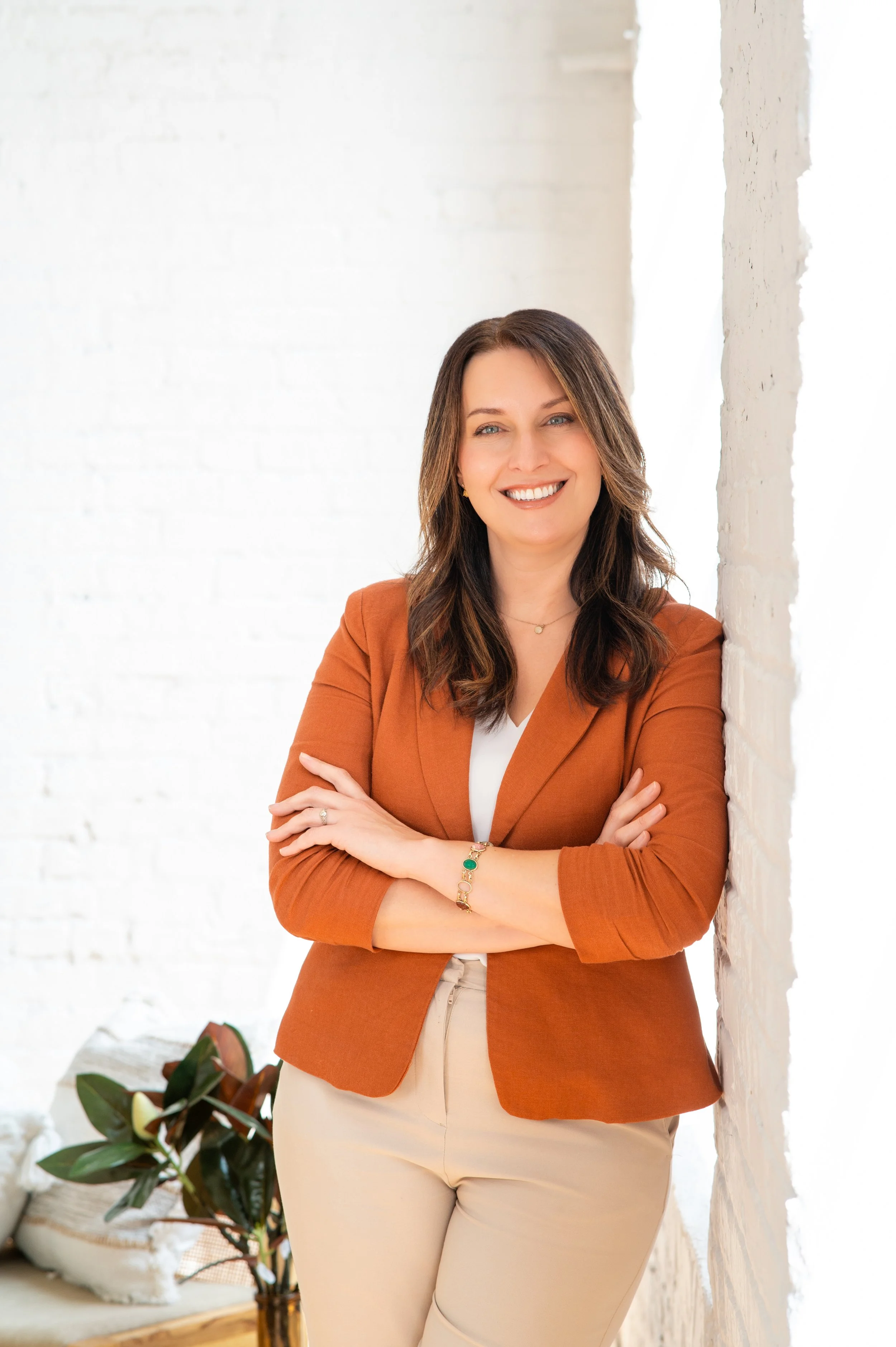 A woman in a rust blazer with her arms folded, against a whitewashed brick wall
