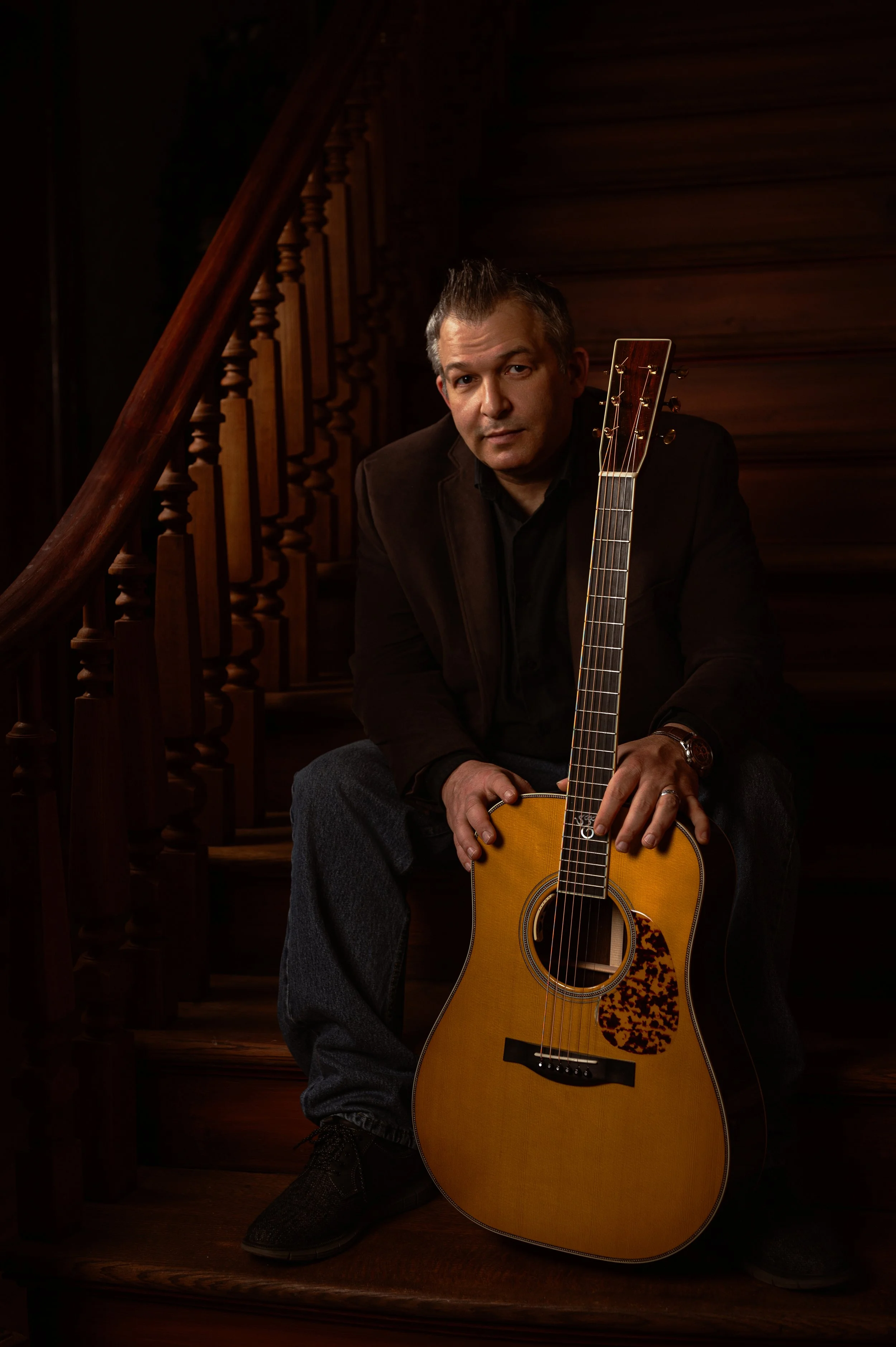 A musician seated on a carved wooden staircase with his acoustic guitar