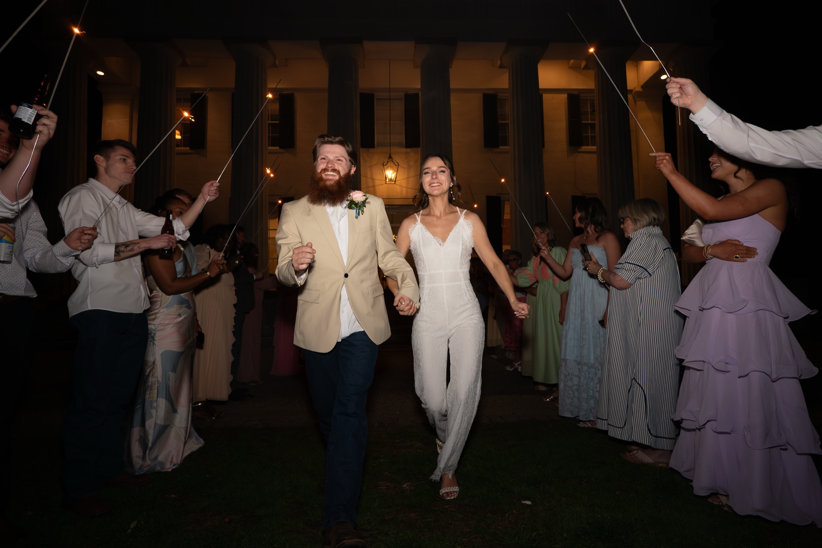 Newlyweds running out through a sparkler send-off in front of a white-columned venue at night