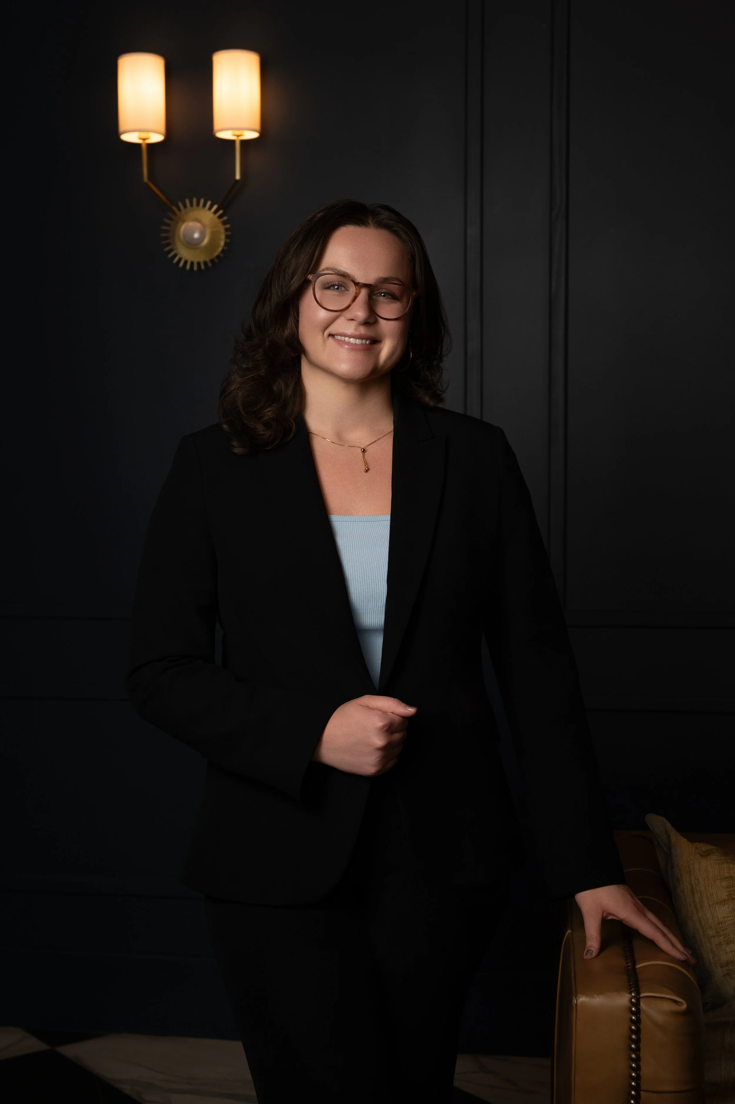 A woman in a black suit and glasses beside a leather chair under a wall sconce