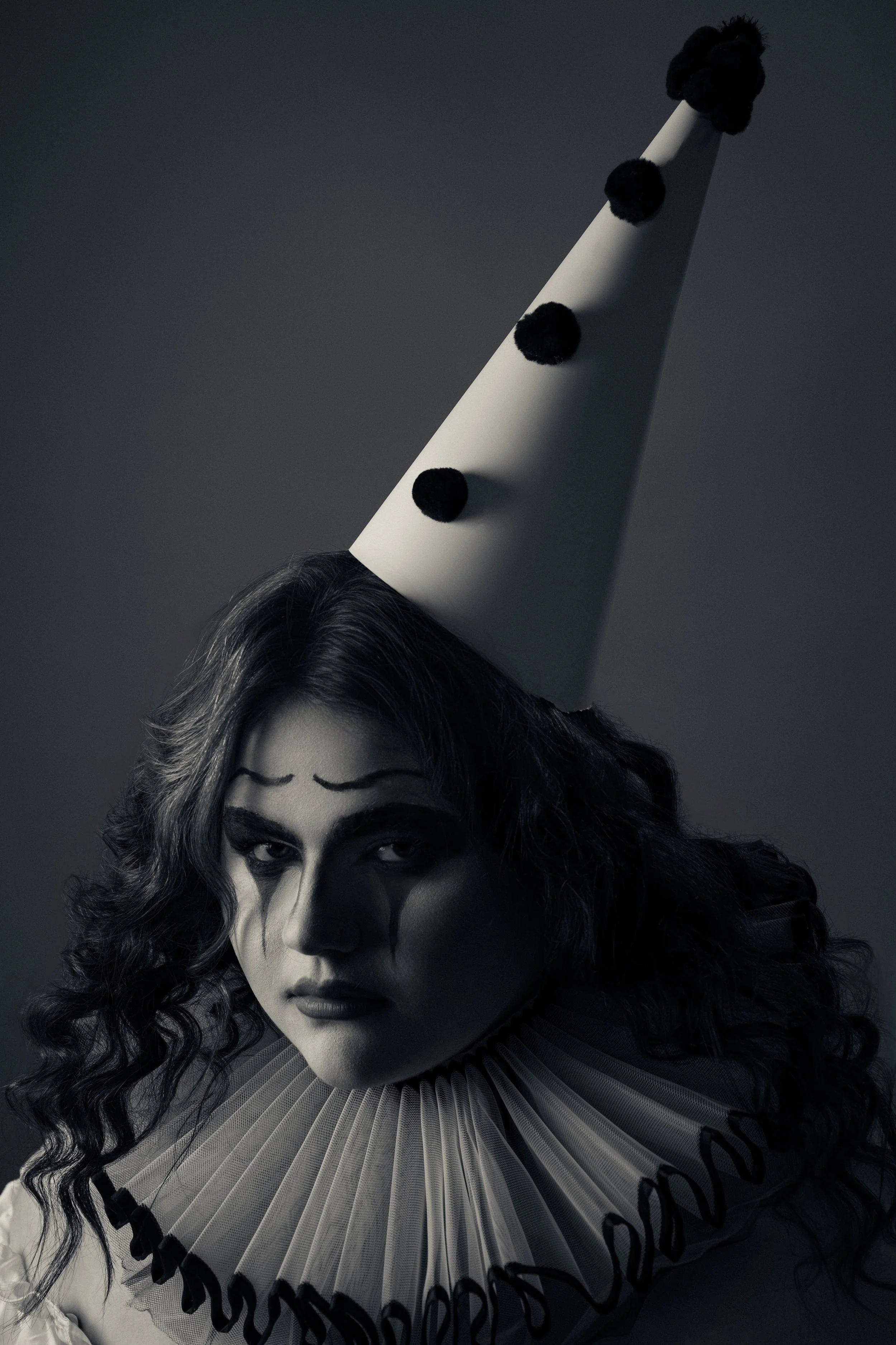 A black-and-white Pierrot portrait — a woman in a ruffled collar and tall pom-pom hat