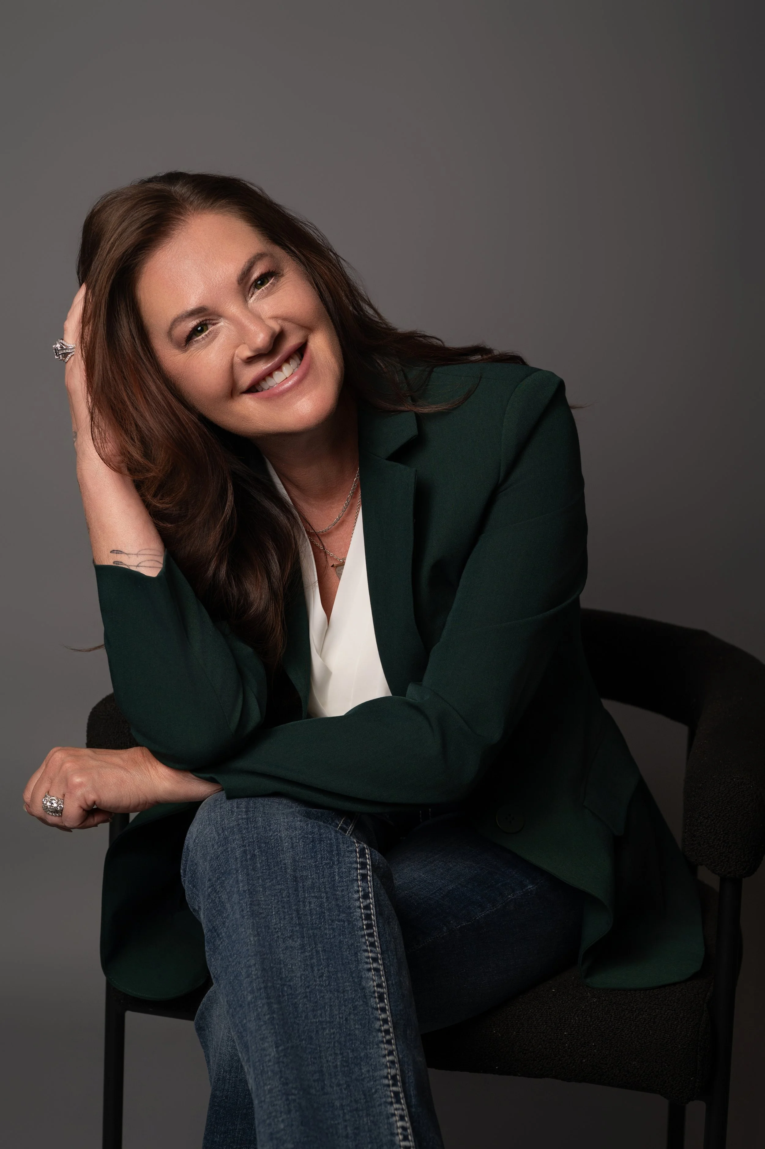 Studio headshot of a smiling professional woman in a dark green blazer against a gray backdrop