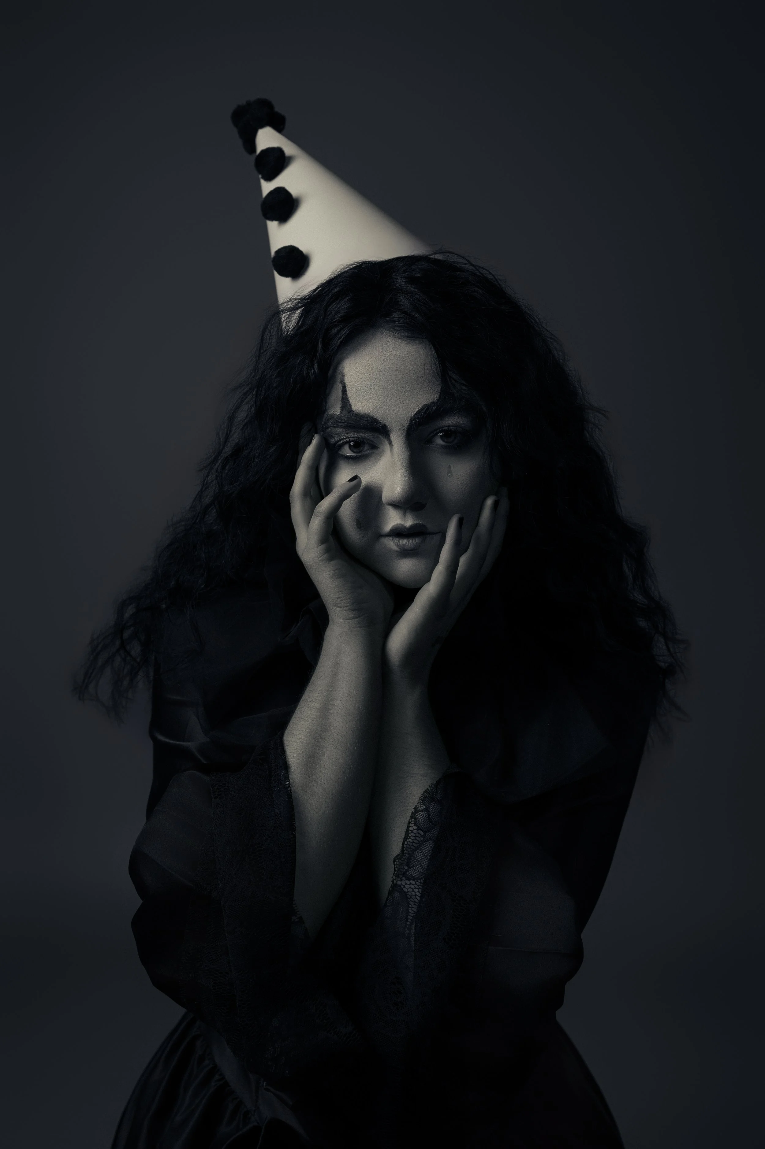 A black-and-white Pierrot portrait — a woman in a pom-pom clown hat resting her face in her hands