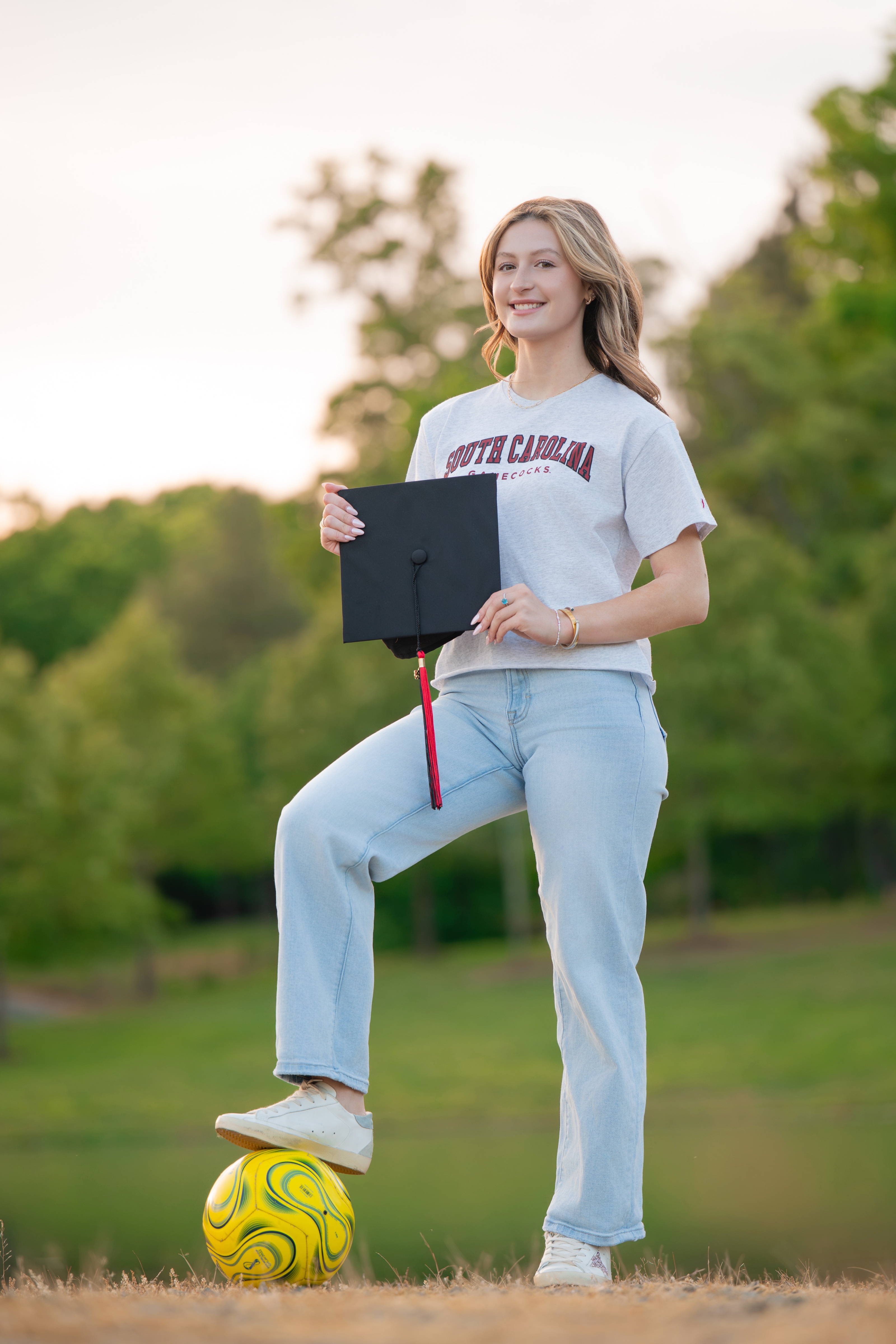 Senior in a college tee balancing a soccer ball underfoot, holding her graduation cap at sunset