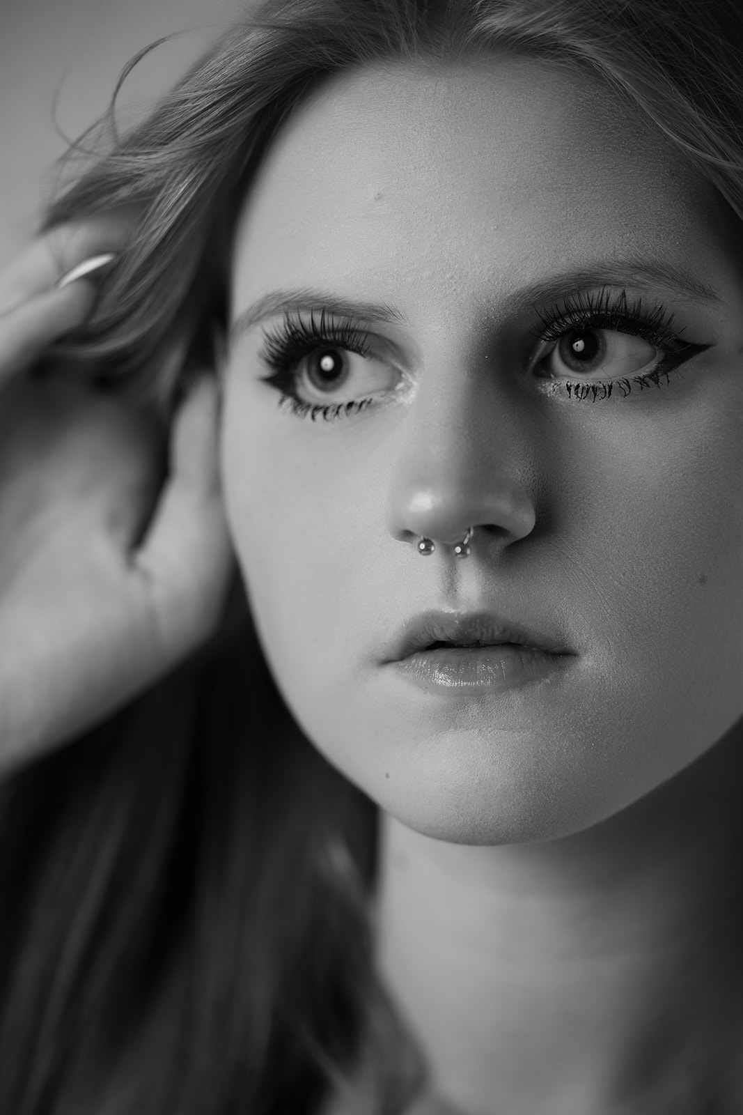 Close-up black-and-white beauty portrait, one hand lifted into her hair, eyes straight down the lens
