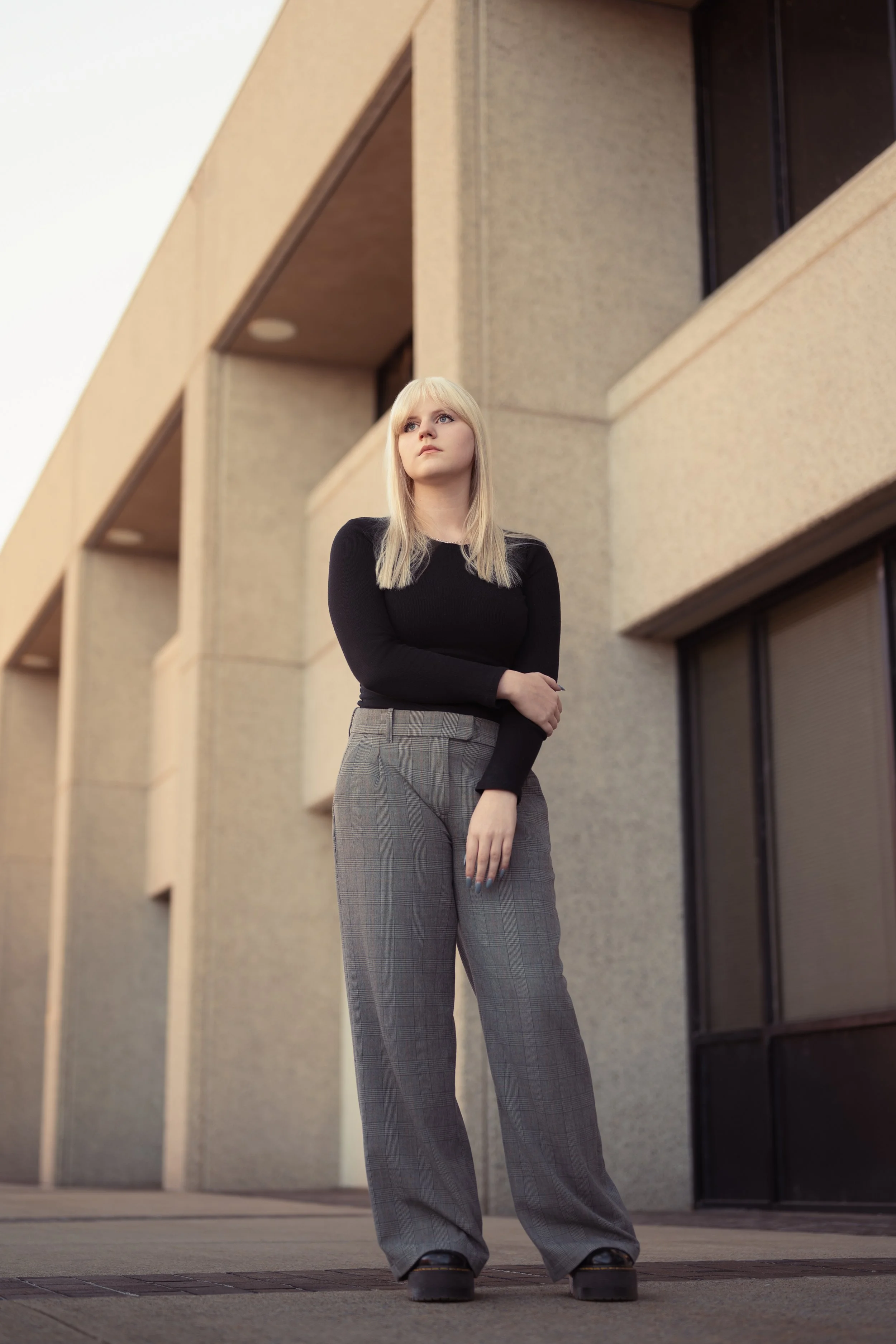 Senior in a black top and plaid trousers standing before a modernist stone building at golden hour