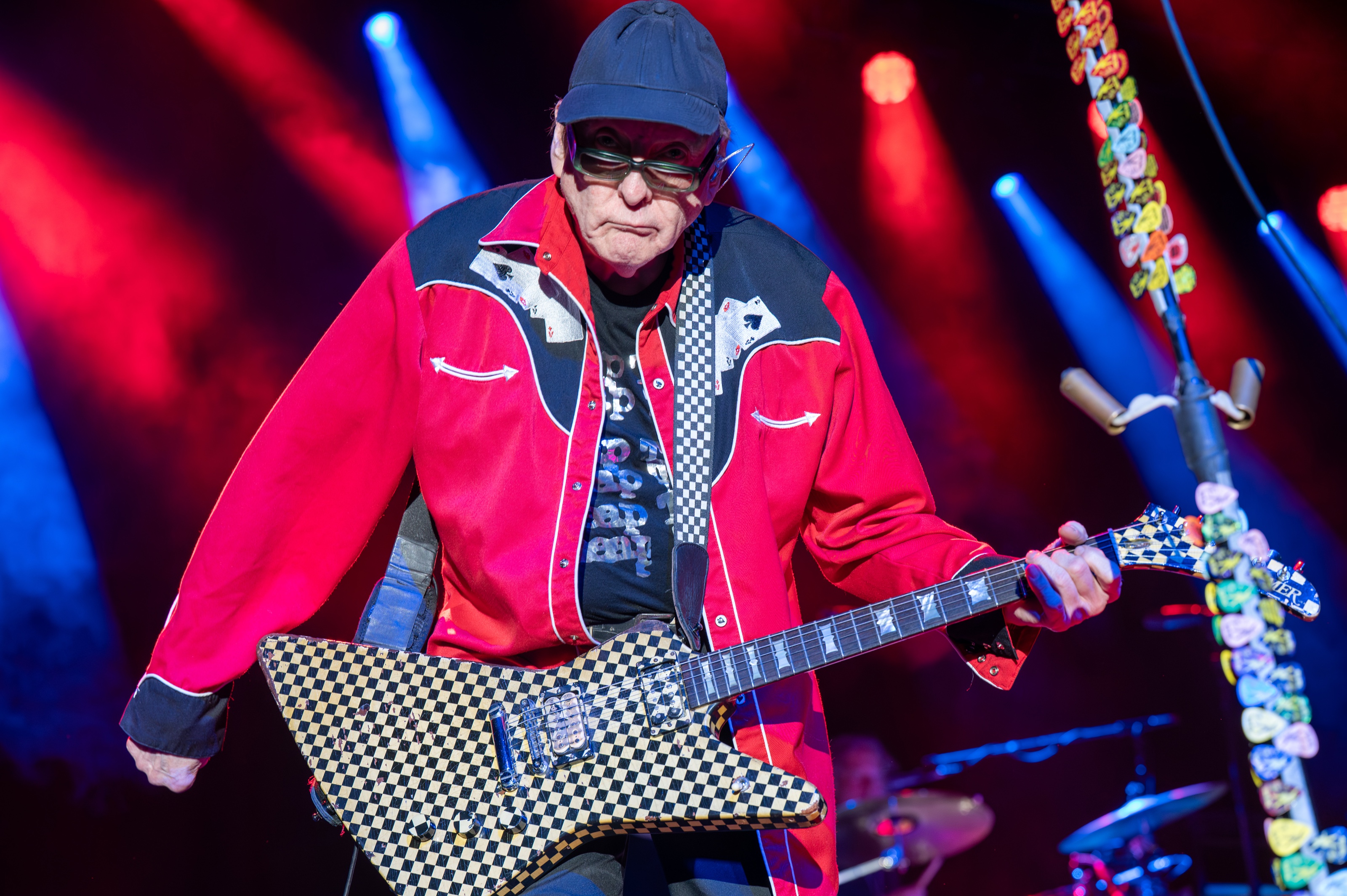 A guitarist in a red western shirt and sunglasses playing a checkerboard electric guitar under red and blue stage light