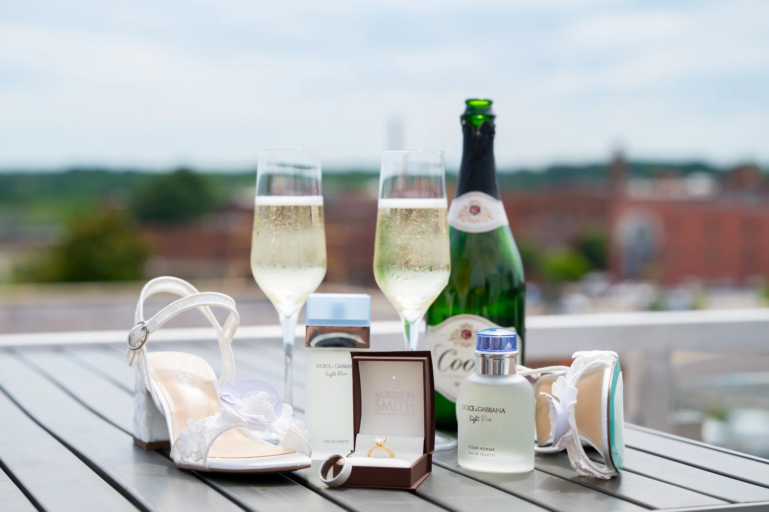 Bridal shoes, perfume, champagne and the ring box arranged on a rooftop table