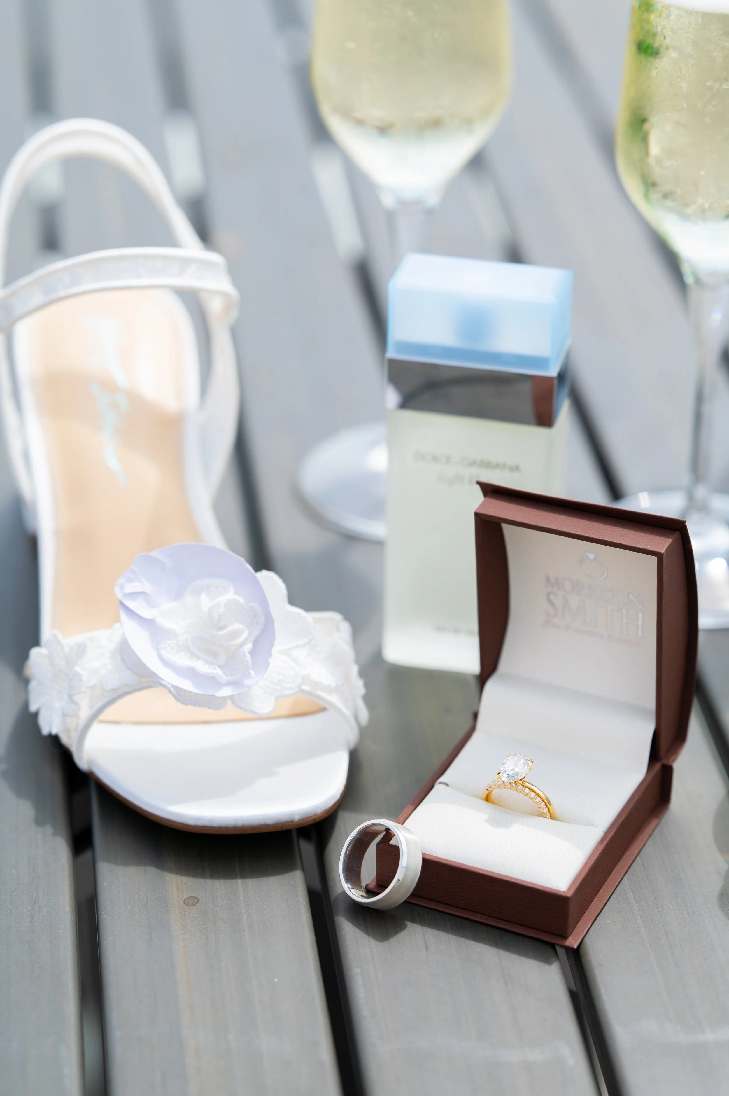 A bridal sandal, the ring in its box, perfume and champagne on a wooden table