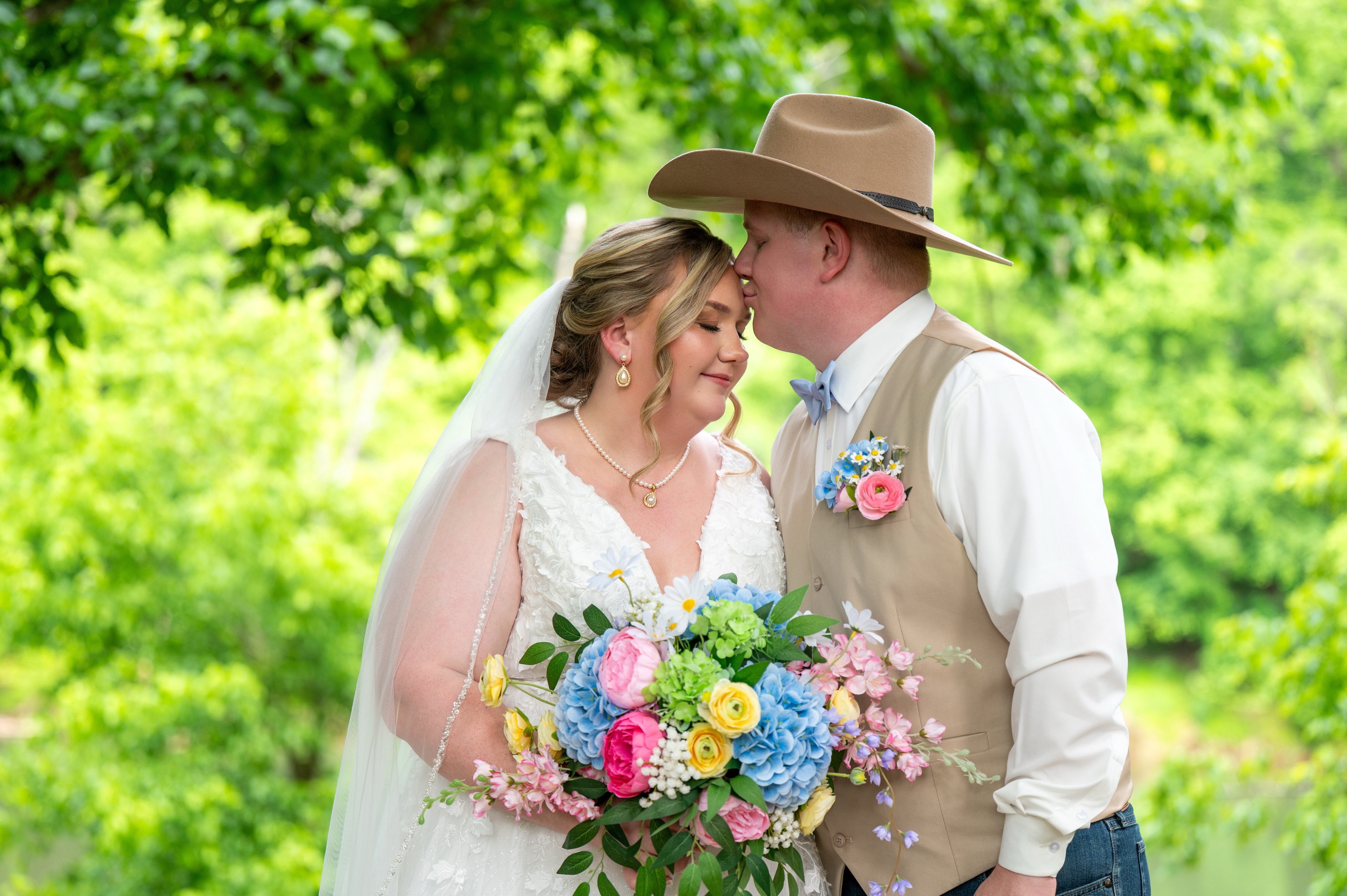 Groom in a cowboy hat kissing his bride's forehead beside a river, her bouquet full of blue and pink blooms