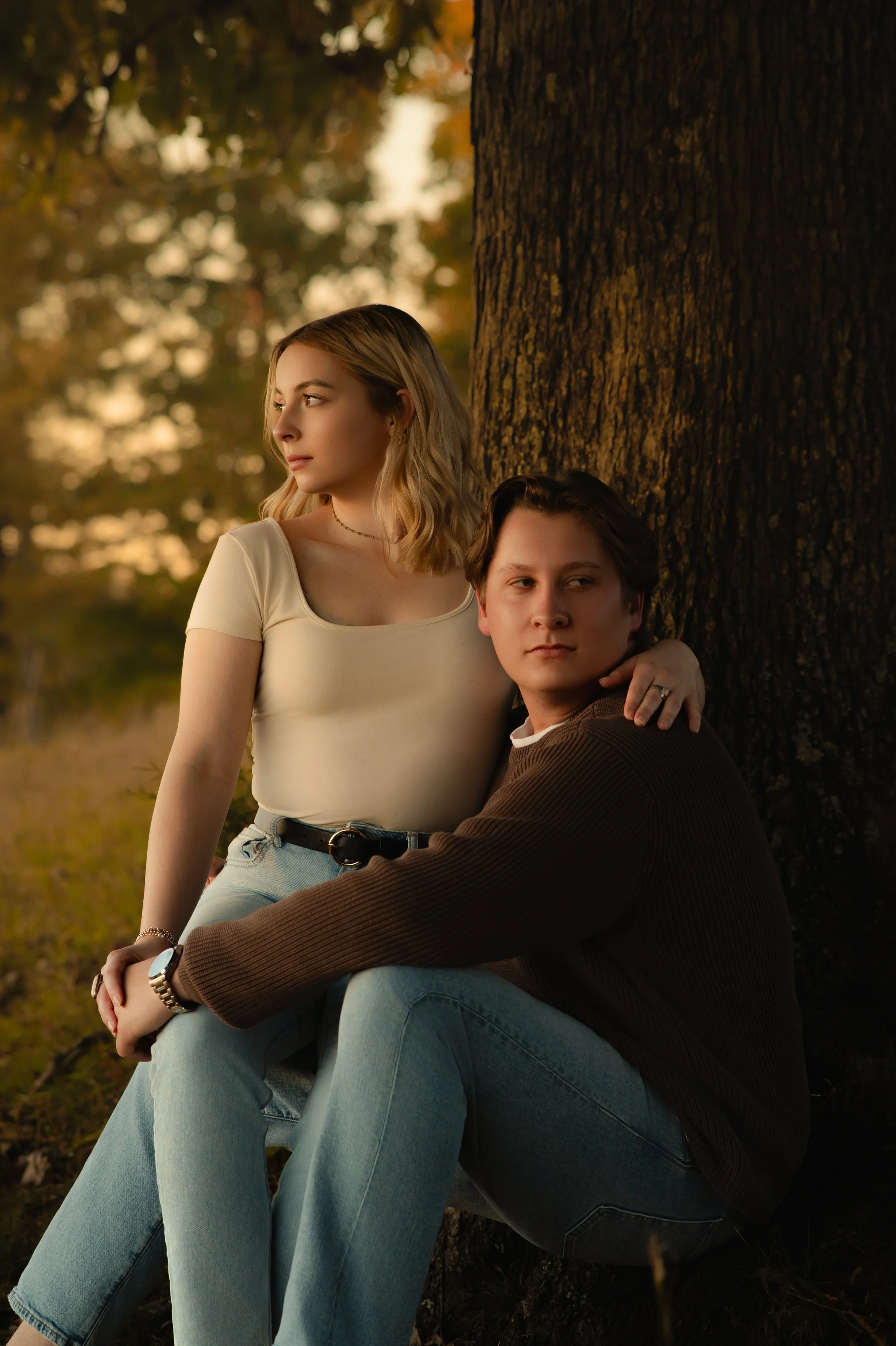 Couple sitting together beneath an oak tree at sunset, her arm around his shoulders