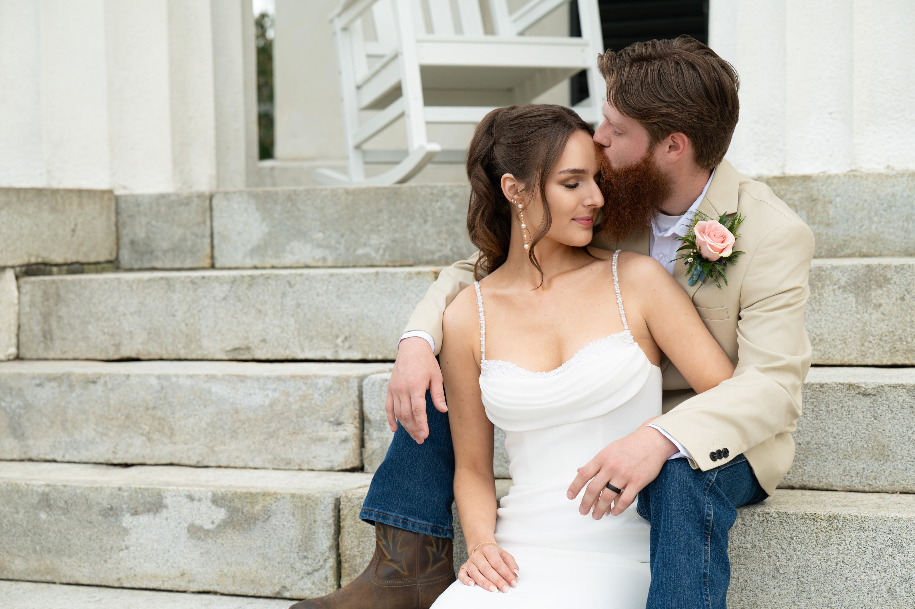 The groom kissing his bride's temple as the two of them sit together on the mansion's stone steps