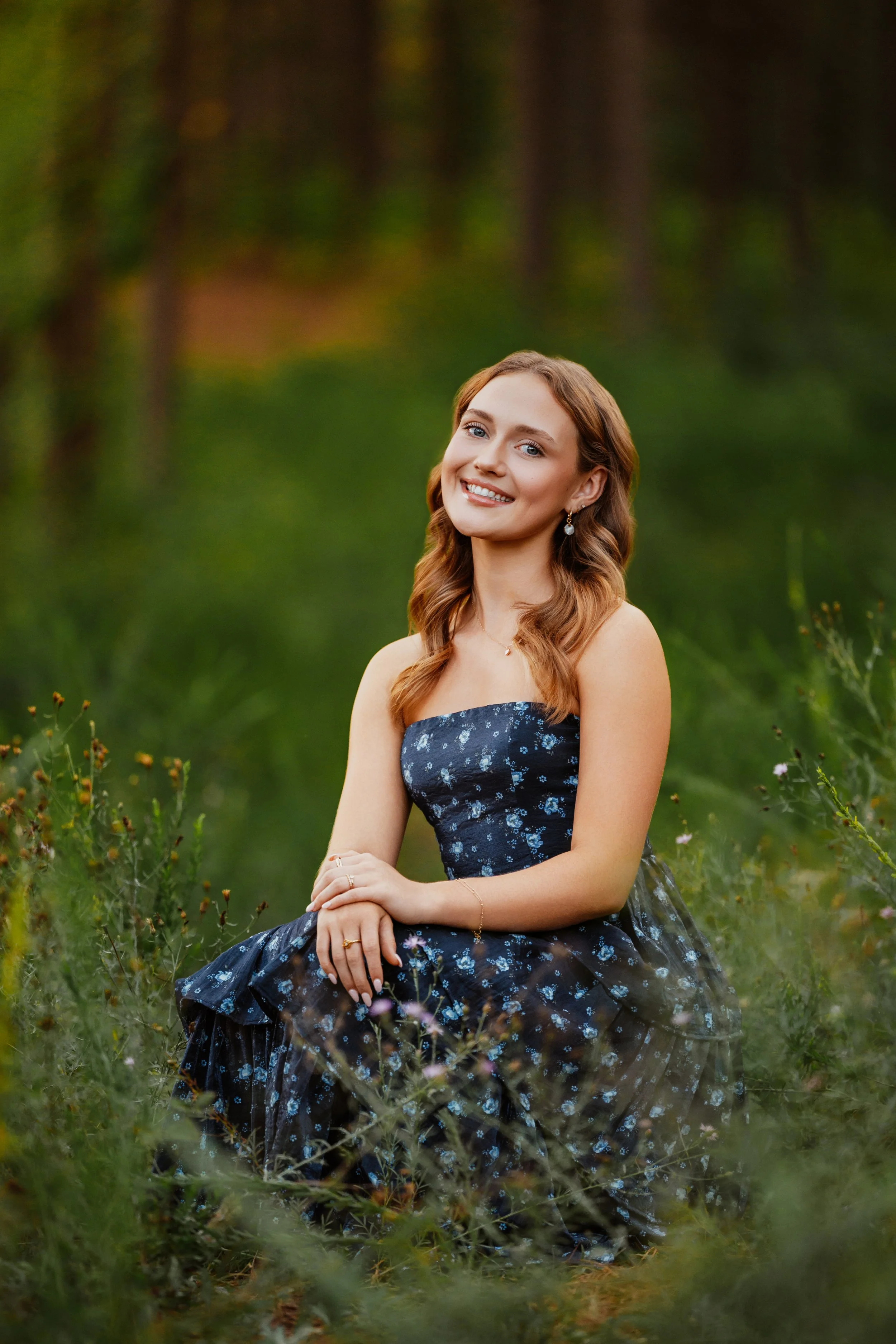 A senior in a navy floral gown seated in tall grass at the edge of the pines