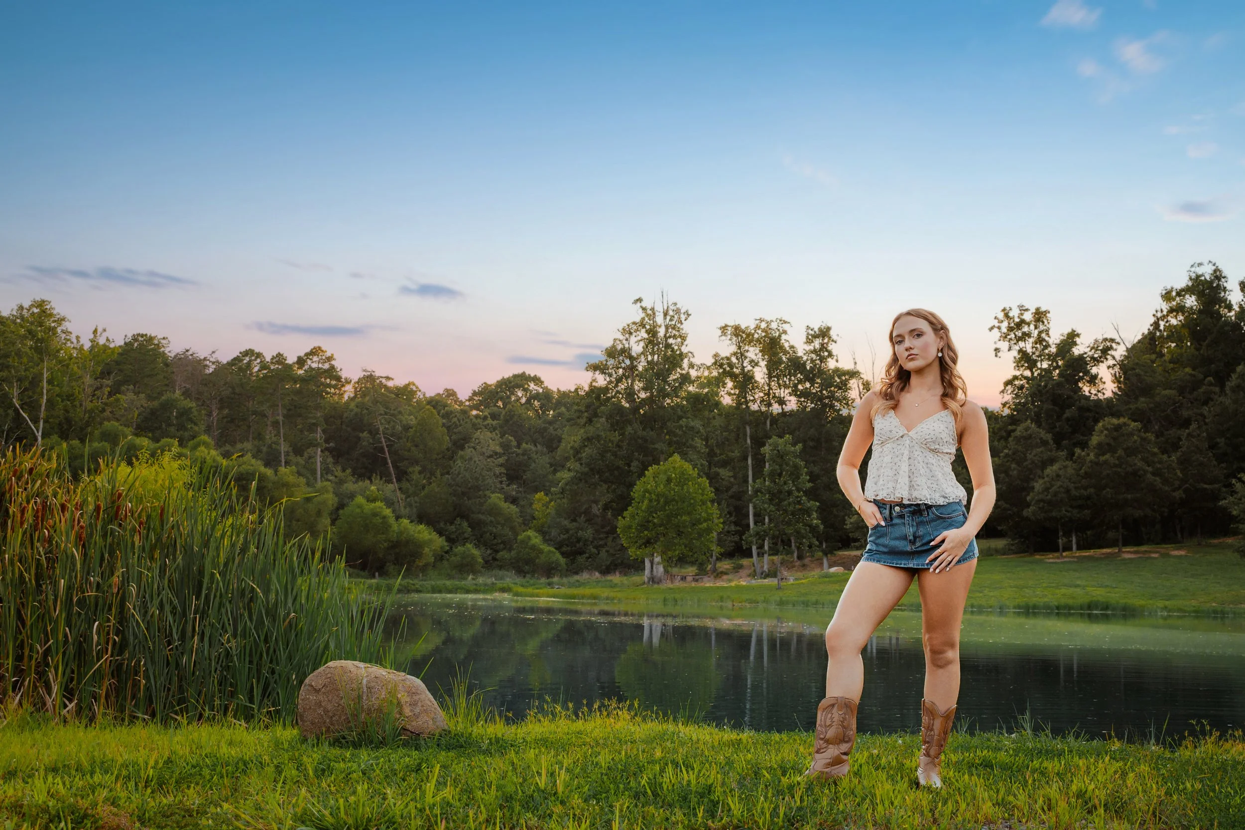 A high school senior in cowboy boots stands beside a pond at sunset — a guided senior portrait session near Danville, Virginia