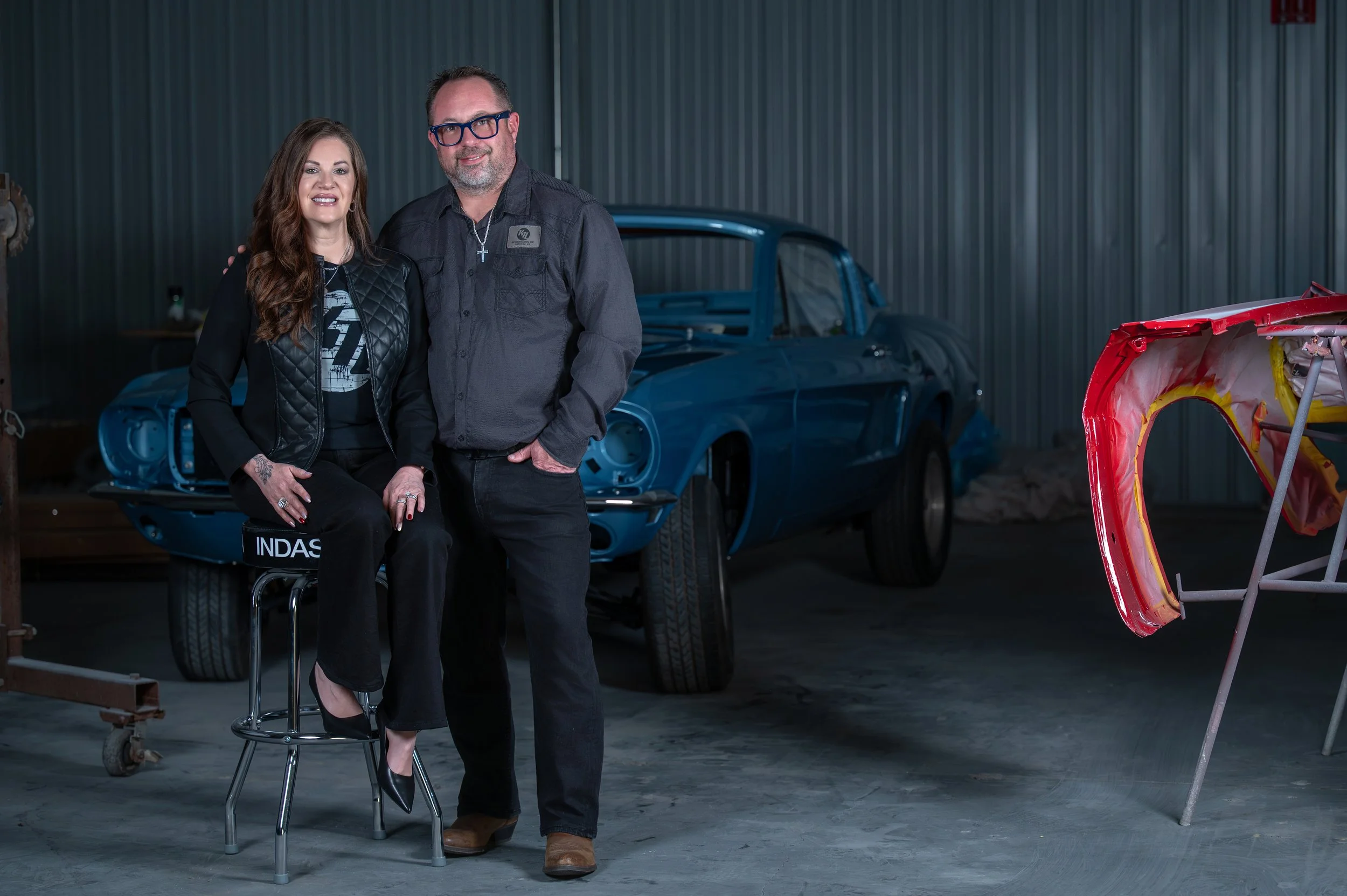 The owners of KTL Restorations photographed magazine-style beside a classic blue Mustang fastback in their Danville restoration shop