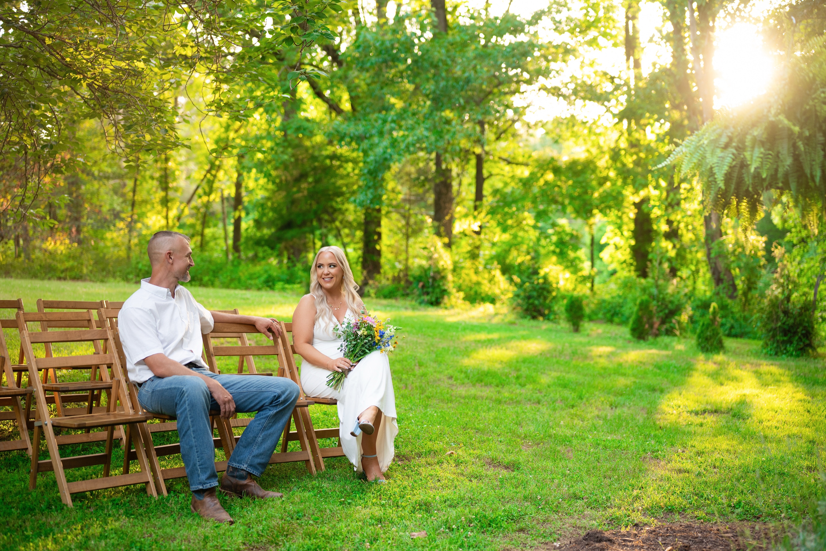 Bride and groom sitting apart on wooden ceremony chairs in a green clearing as the sun breaks through the trees