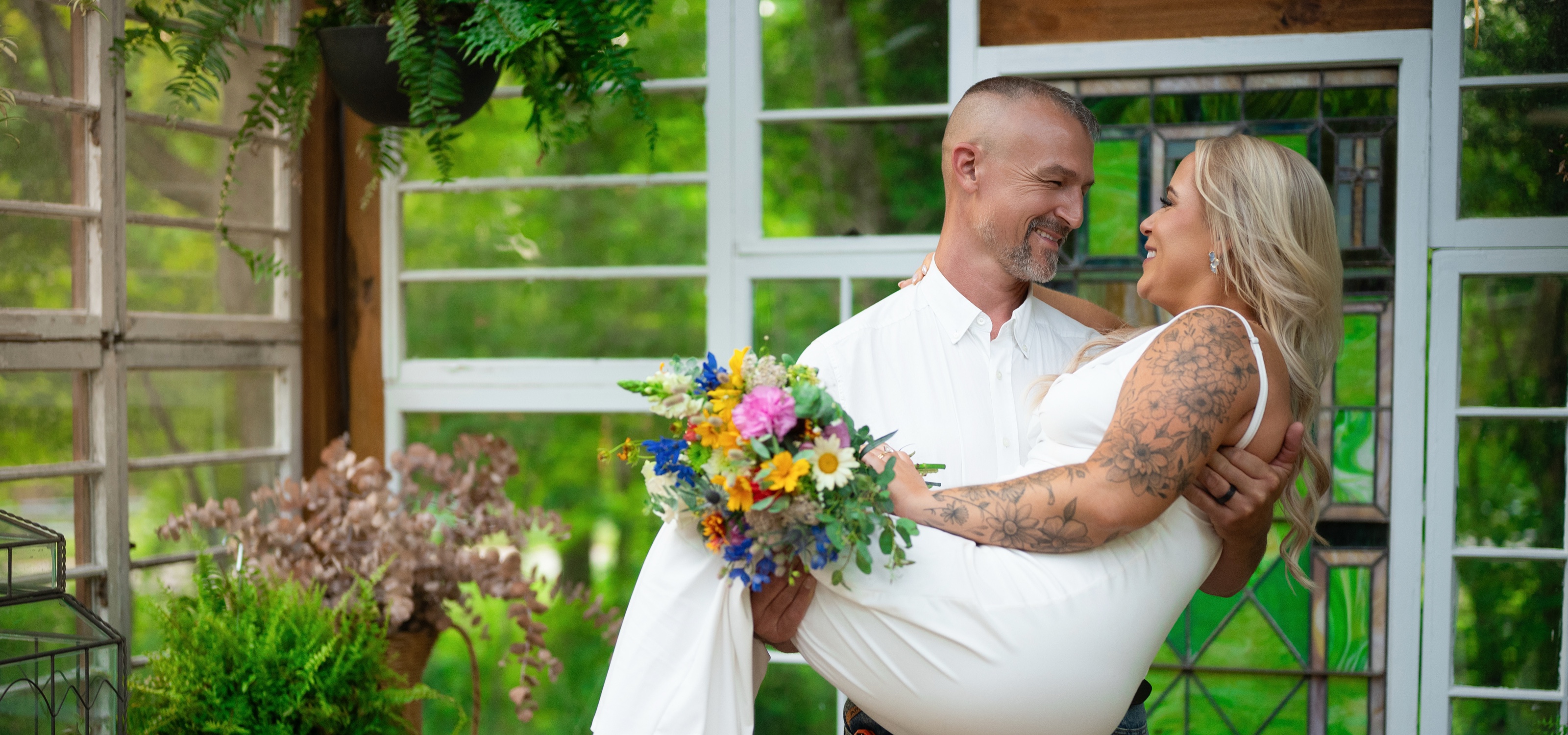 Groom carrying his laughing bride through a greenhouse doorway, her bouquet of wildflowers between them
