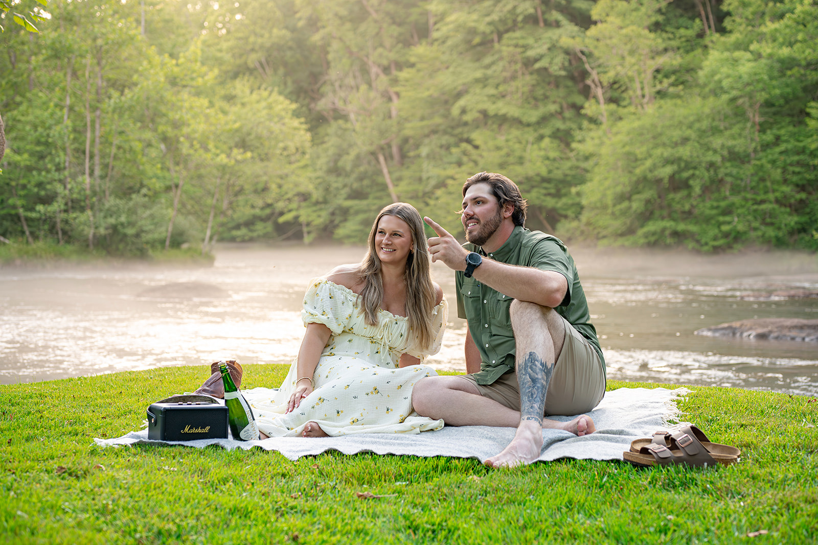 Couple sitting on a picnic blanket beside a misty river at golden hour with a speaker and champagne