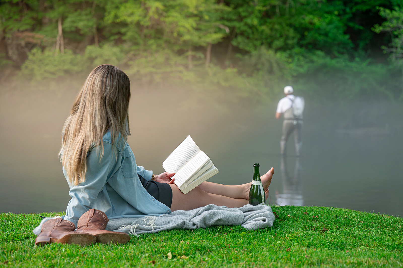 Woman reading on a riverbank blanket while a fly fisherman casts in the mist behind her