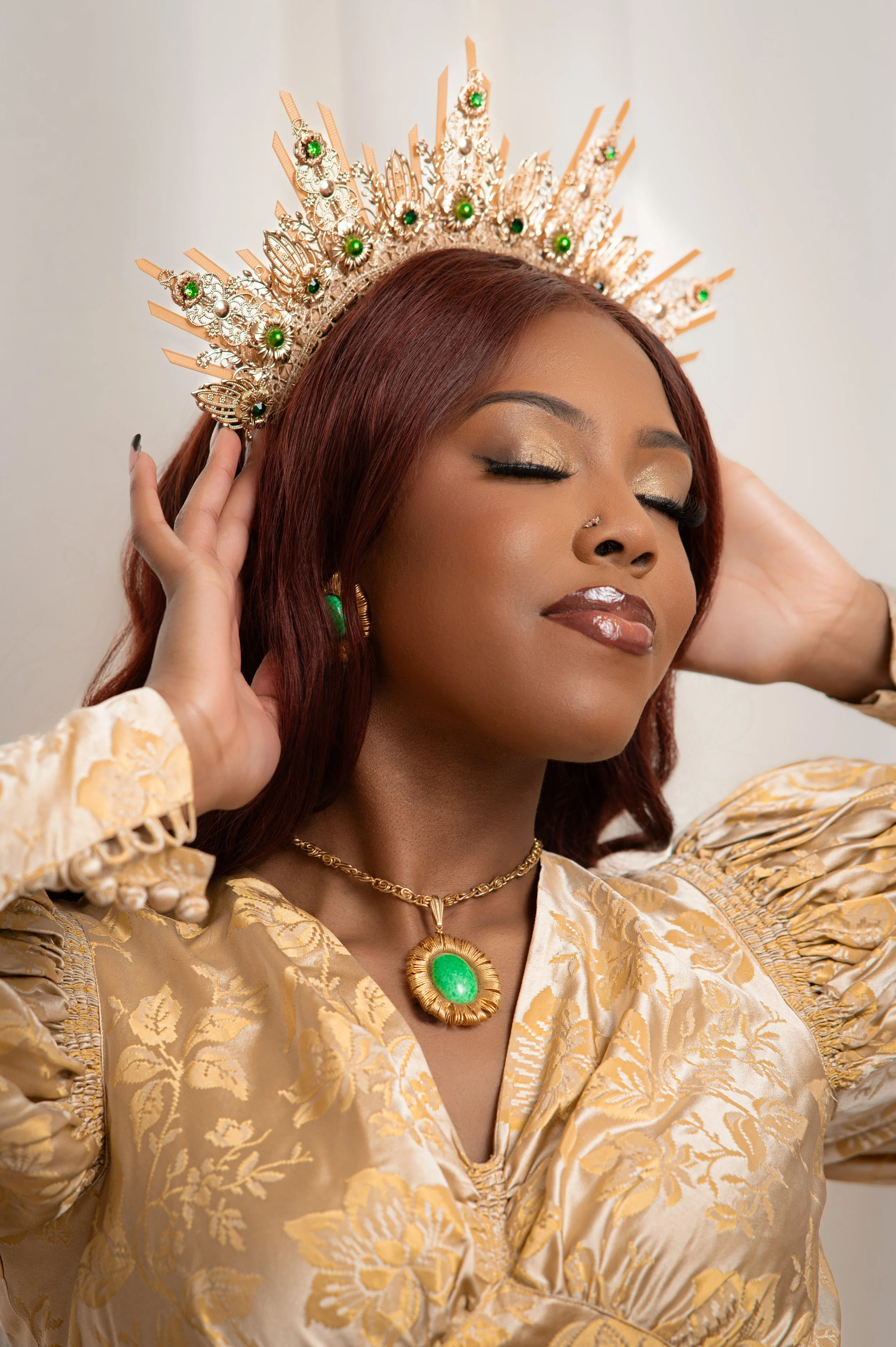 Editorial studio portrait of a senior in a gold brocade gown and jeweled crown, eyes closed under soft light