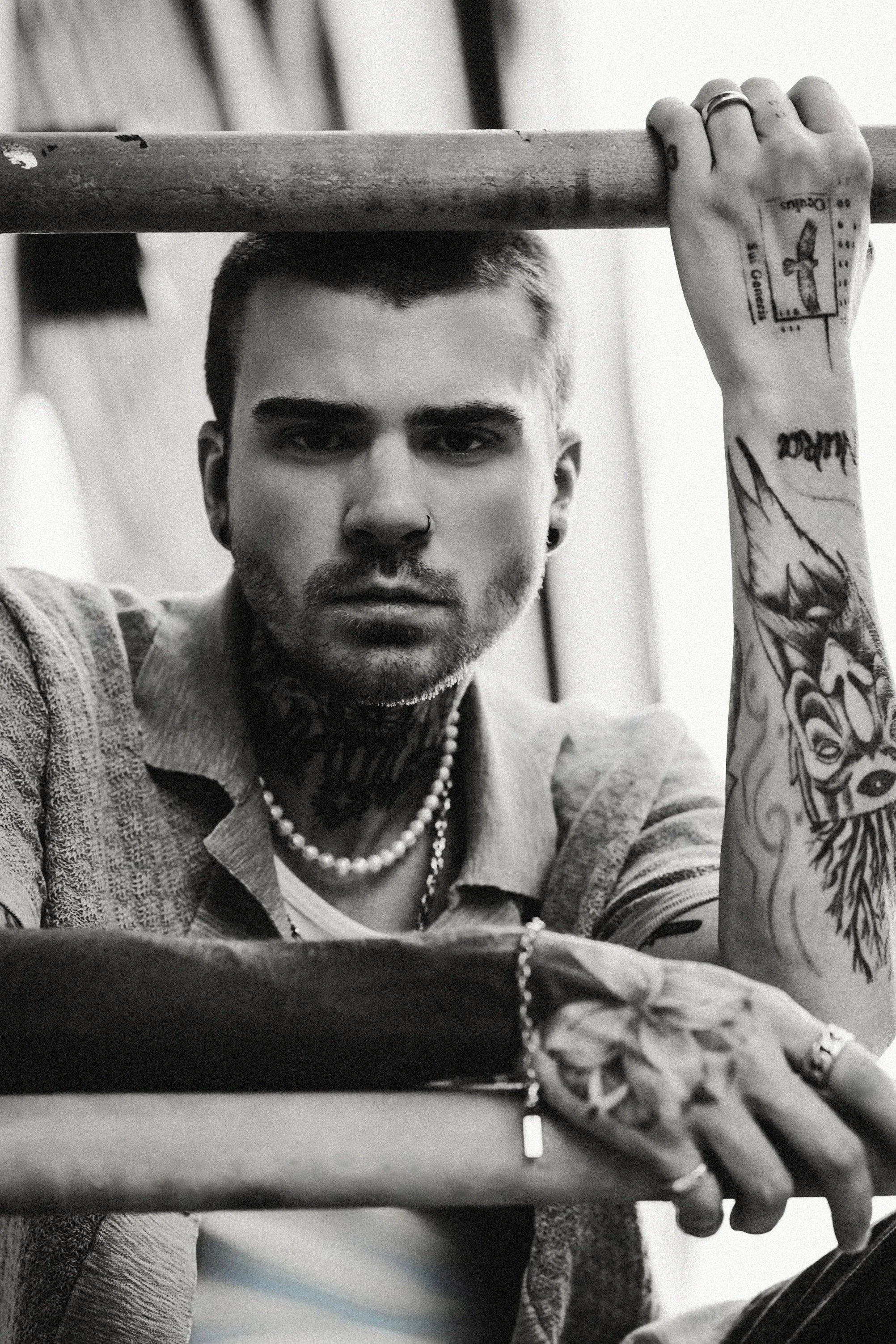 Patrick Gibson — black-and-white self-portrait, tattooed arms resting on a metal rail, looking straight into the camera