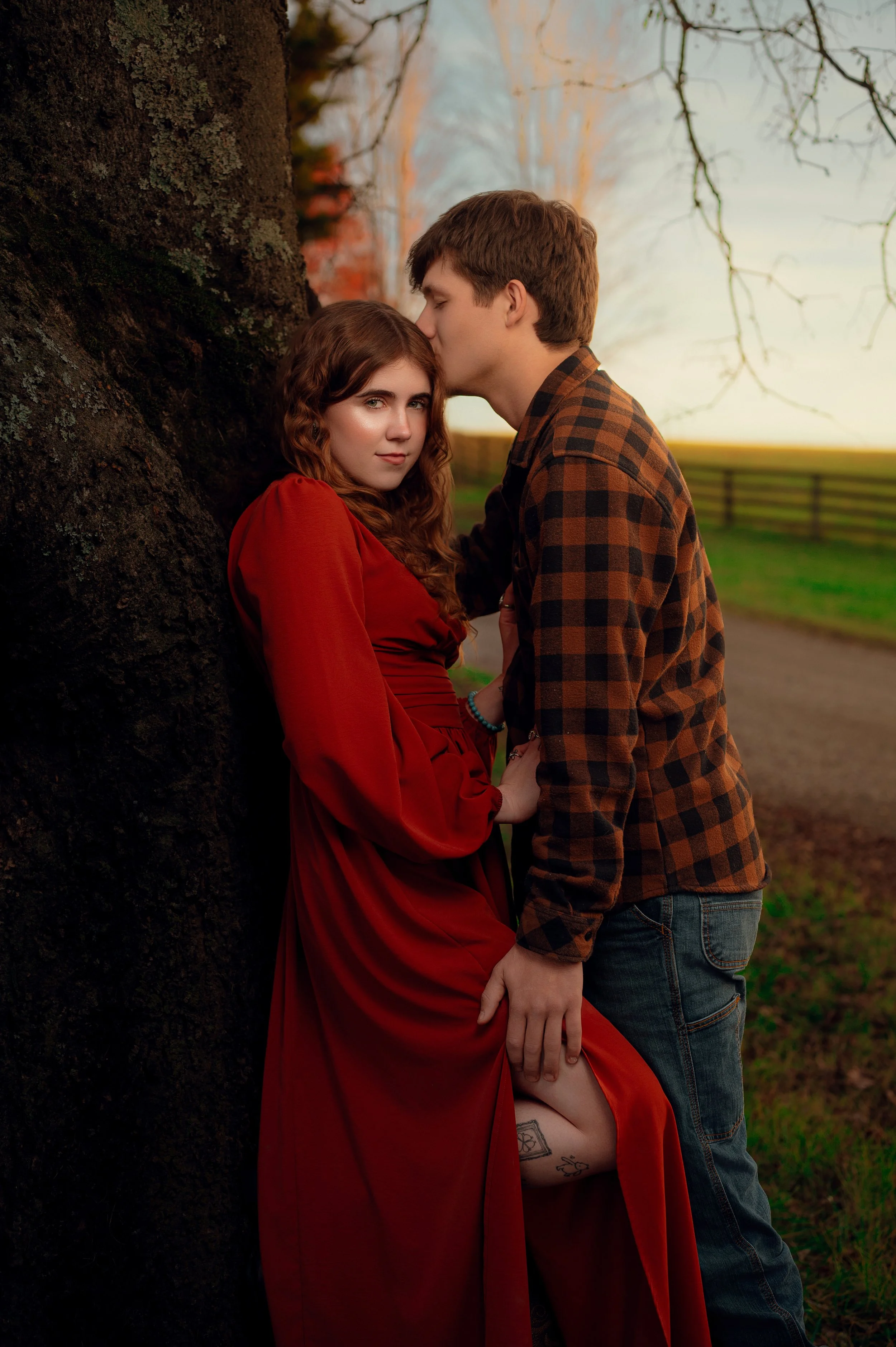 Man in a plaid flannel kissing the forehead of a woman in a long red dress beside a farm lane at sunset
