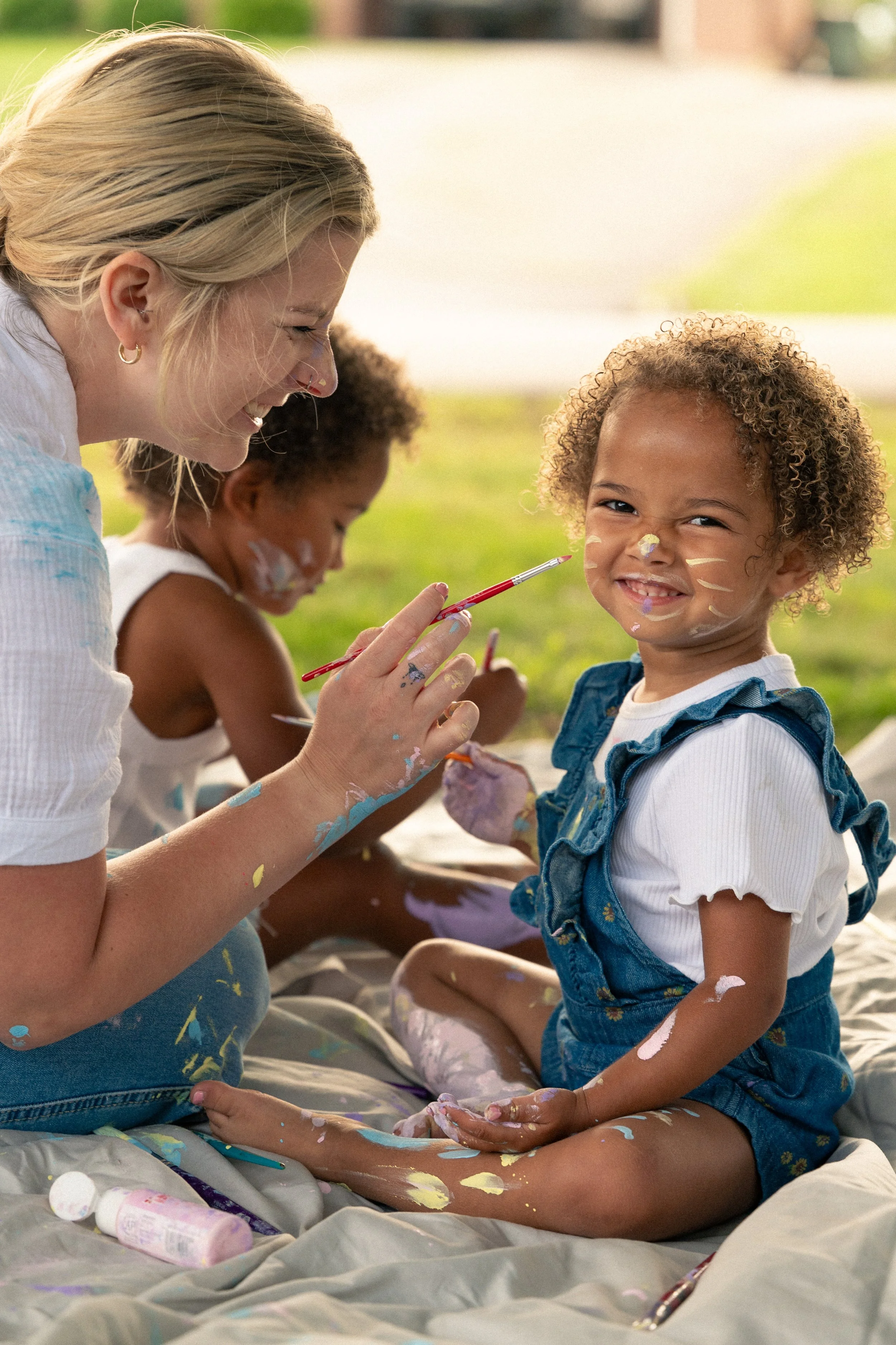 Mom laughing while painting her giggling daughter's face during a backyard paint session