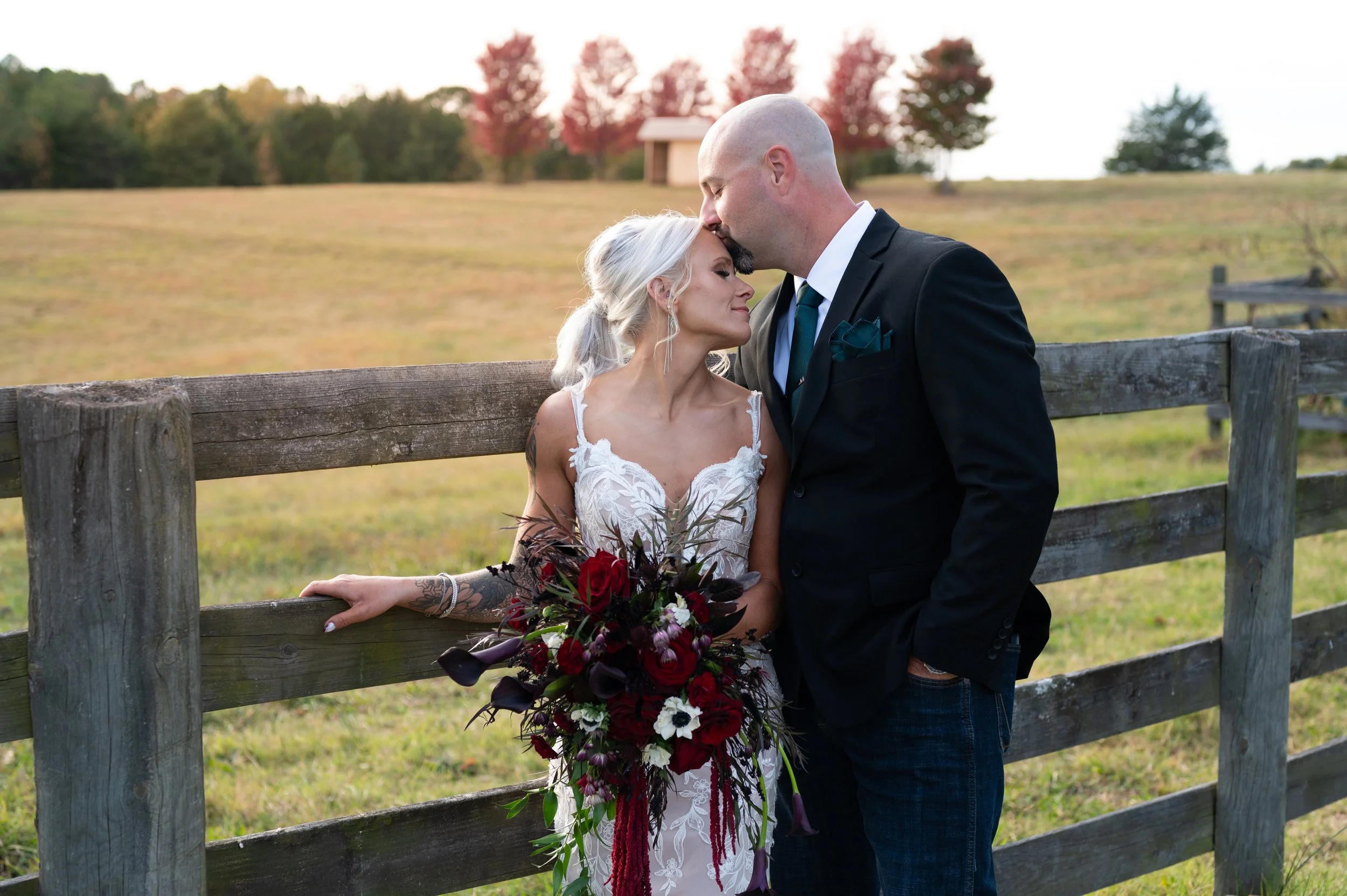 A groom kissing his bride's forehead at golden hour beside a wooden fence