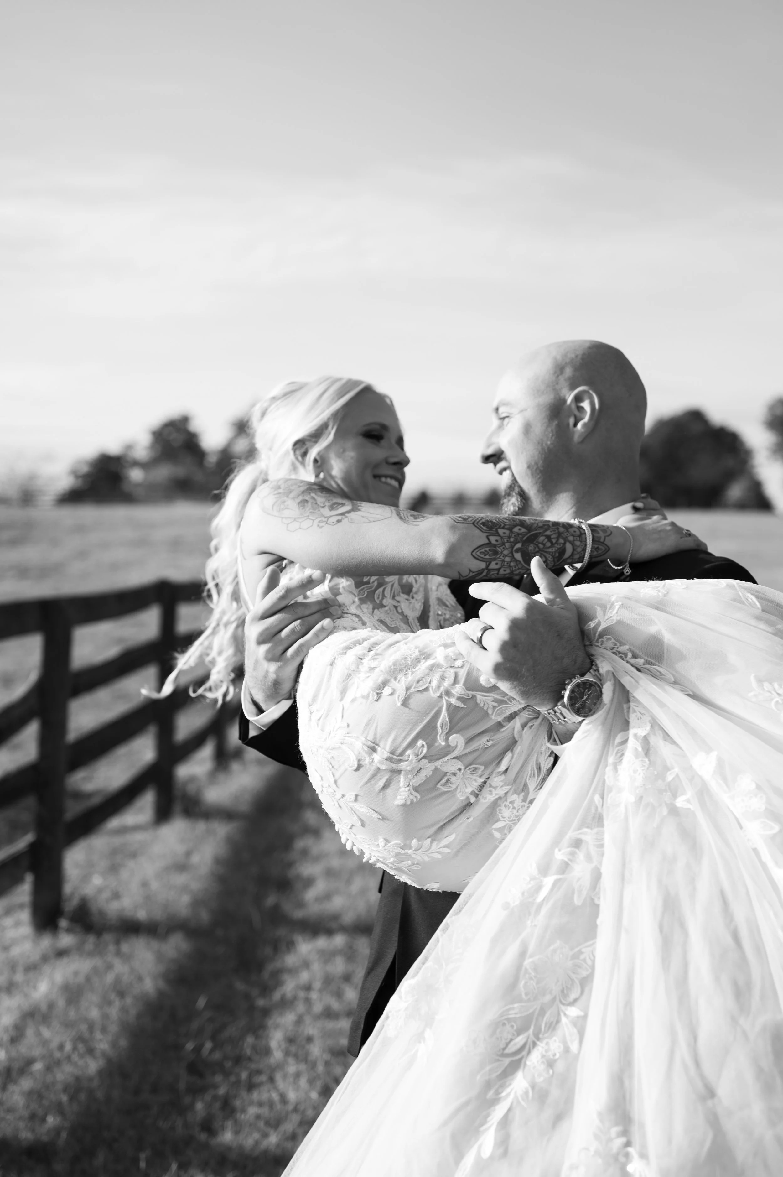 Bald, bearded groom in a dark suit carrying his bride along a wooden farm fence at golden hour, in black and white