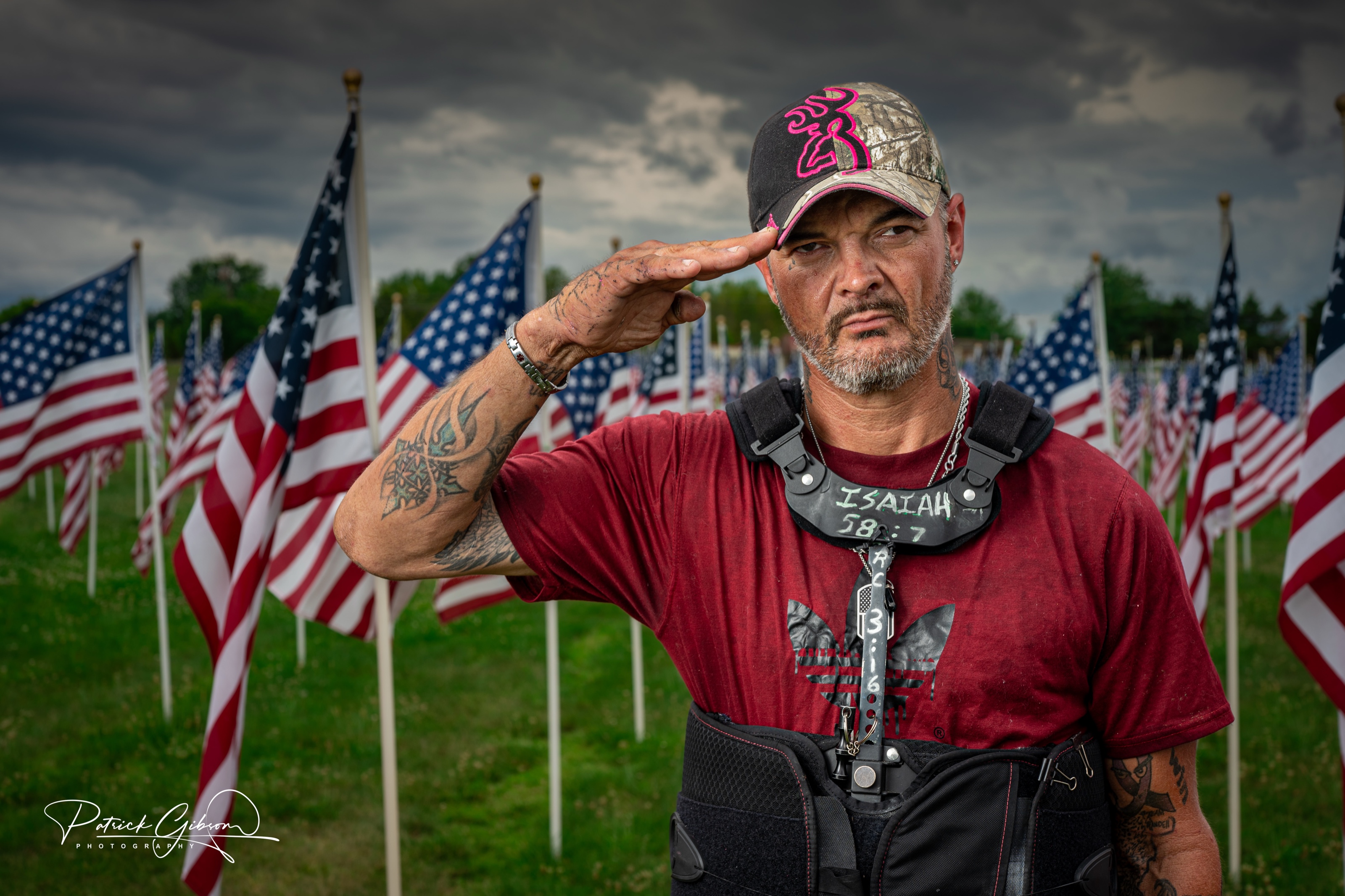 A veteran in a ball cap saluting in front of hundreds of American flags under a storm sky