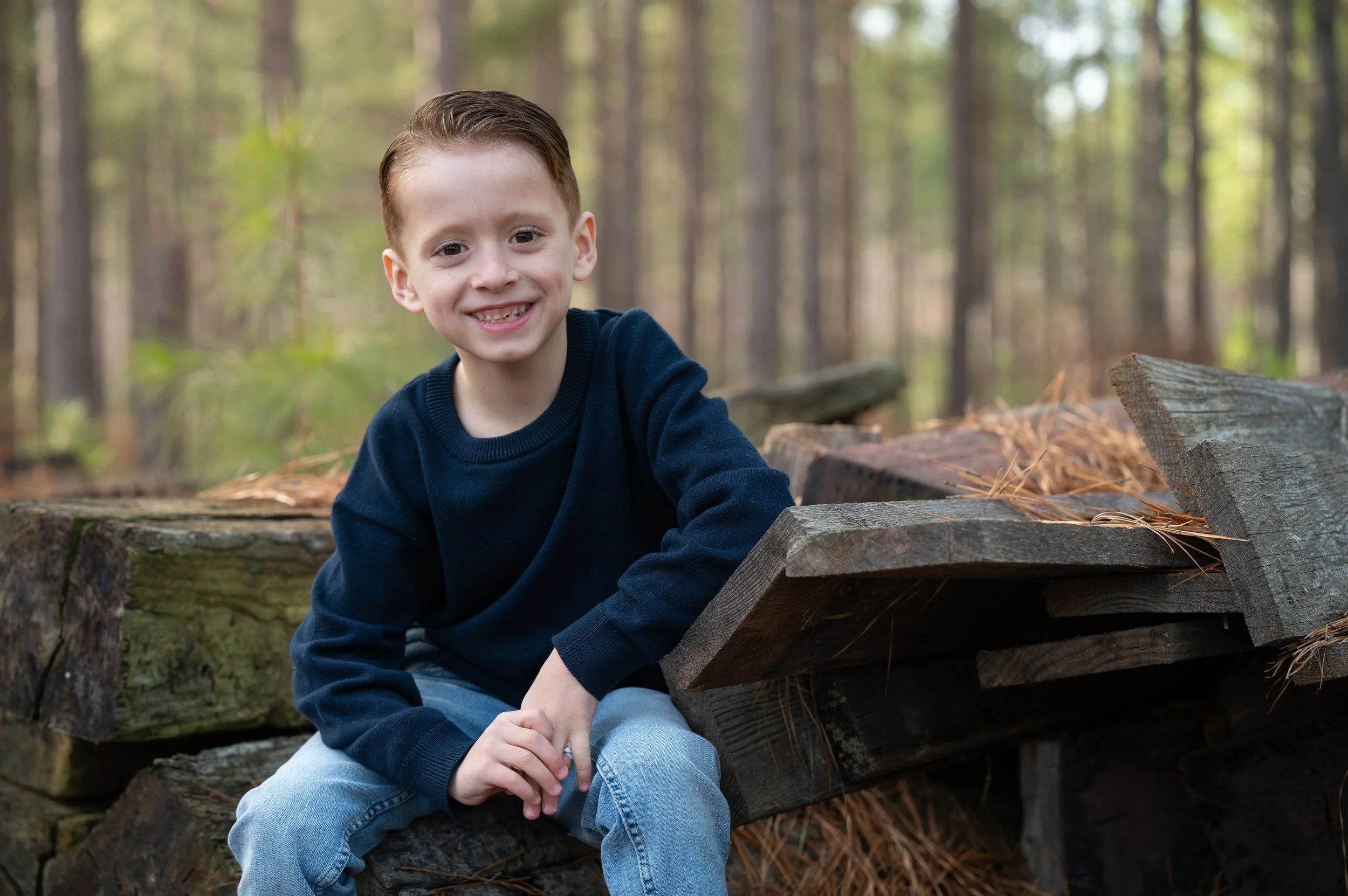Young boy in a navy sweater smiling from a stack of weathered timbers in a pine forest