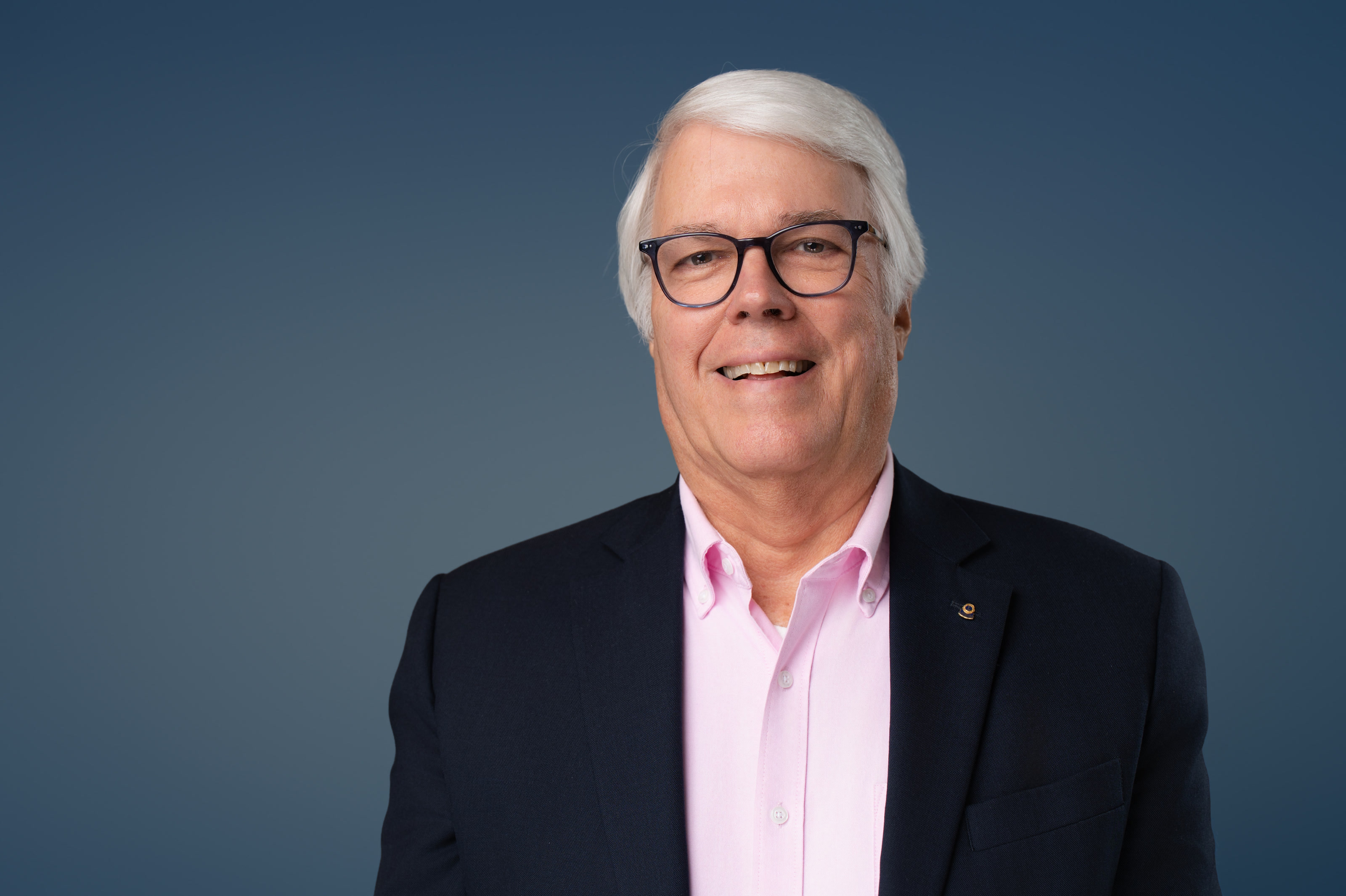 Studio headshot of a smiling man with white hair, glasses and a navy jacket against a deep blue backdrop
