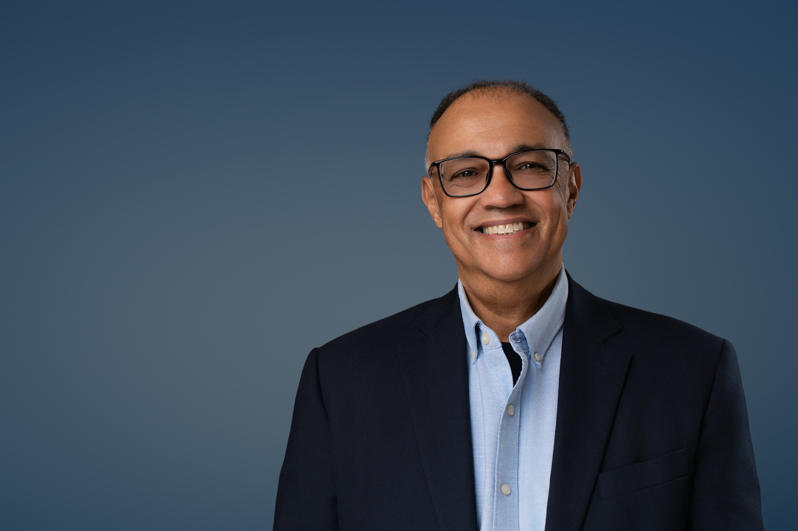Studio headshot of a smiling man in a navy suit and glasses against a deep blue backdrop