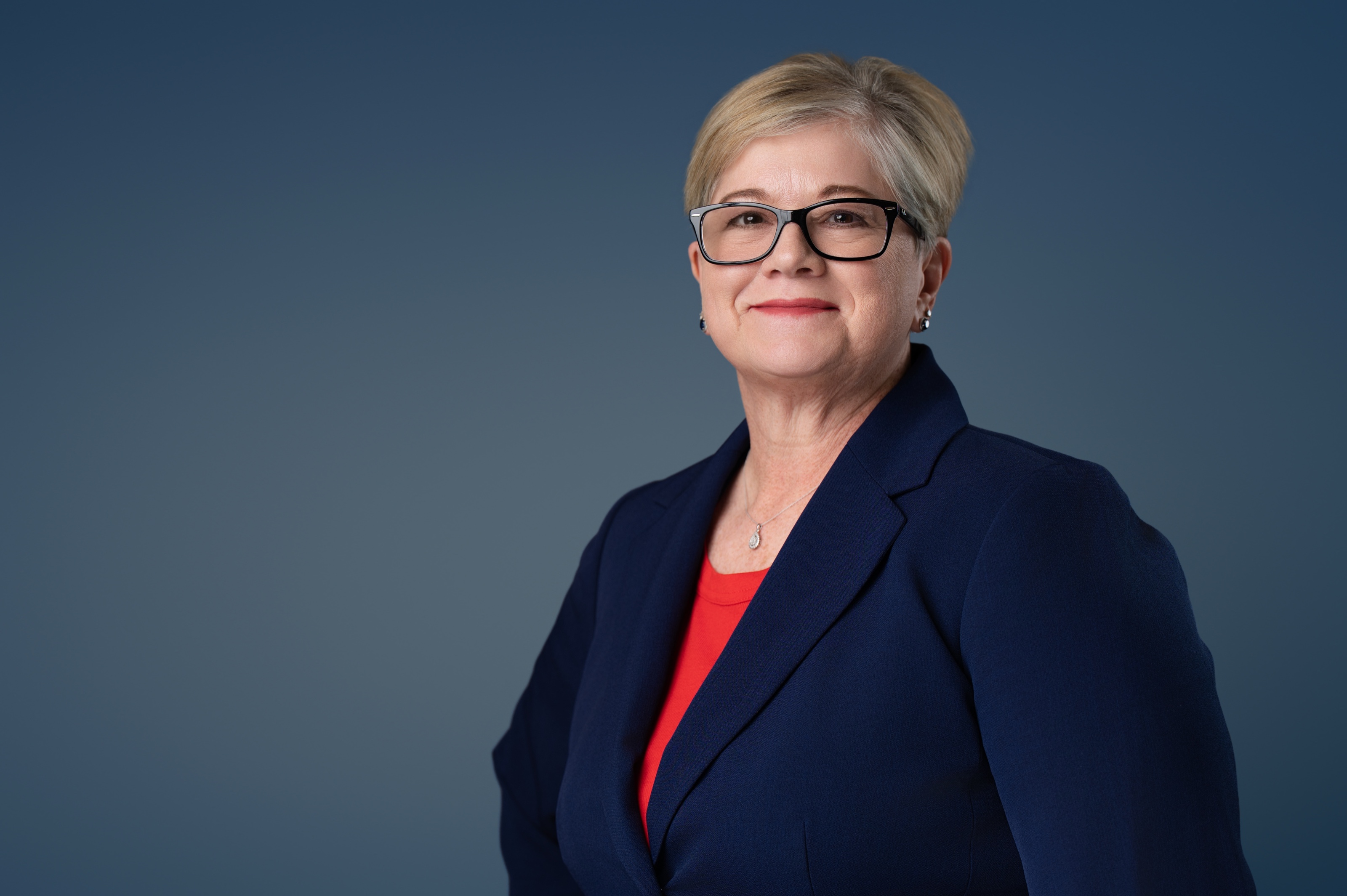 Studio headshot of a woman in a navy blazer over a red top against a deep blue backdrop