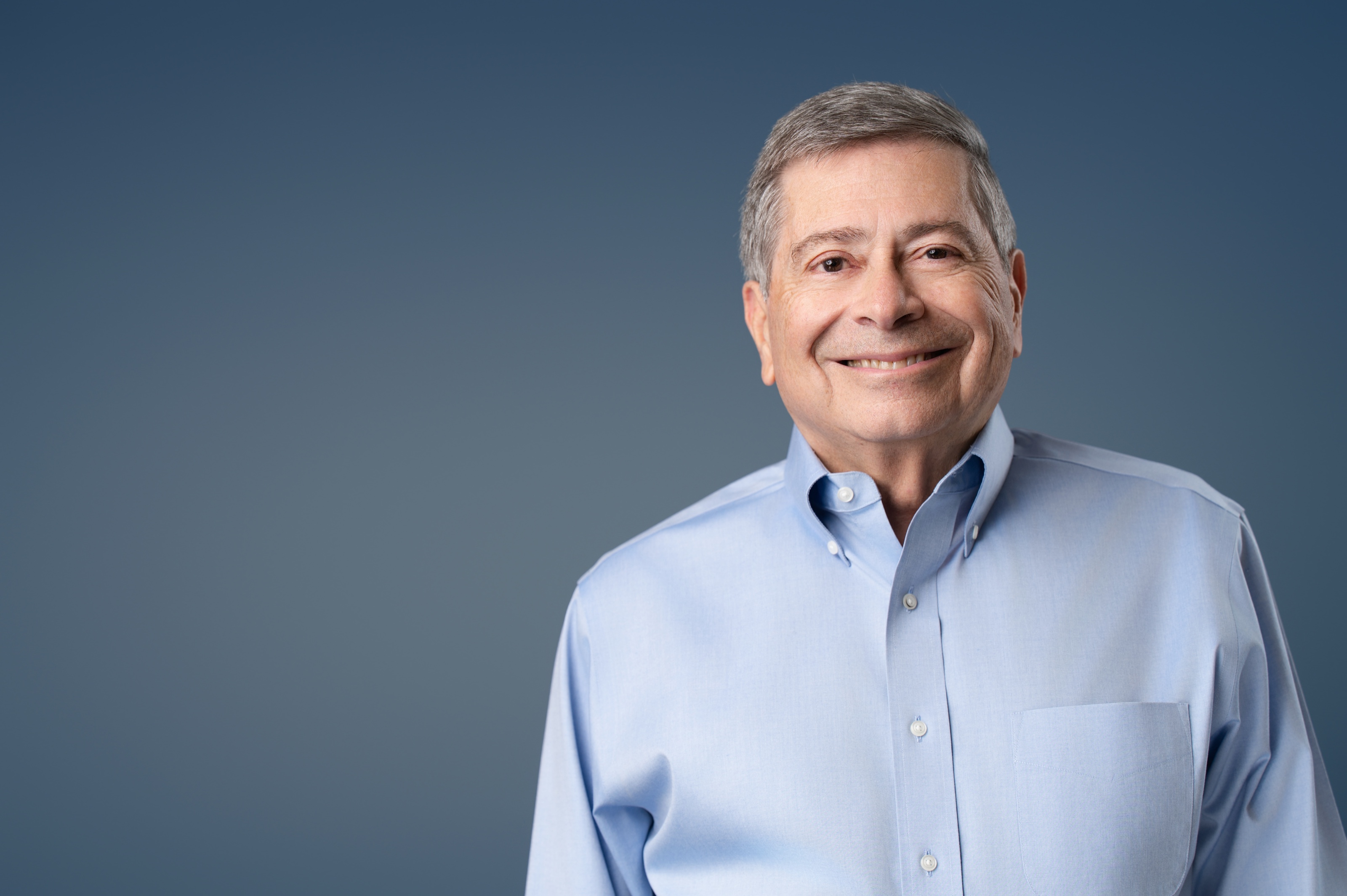 Studio headshot of a smiling man in a light blue oxford shirt against a deep blue backdrop