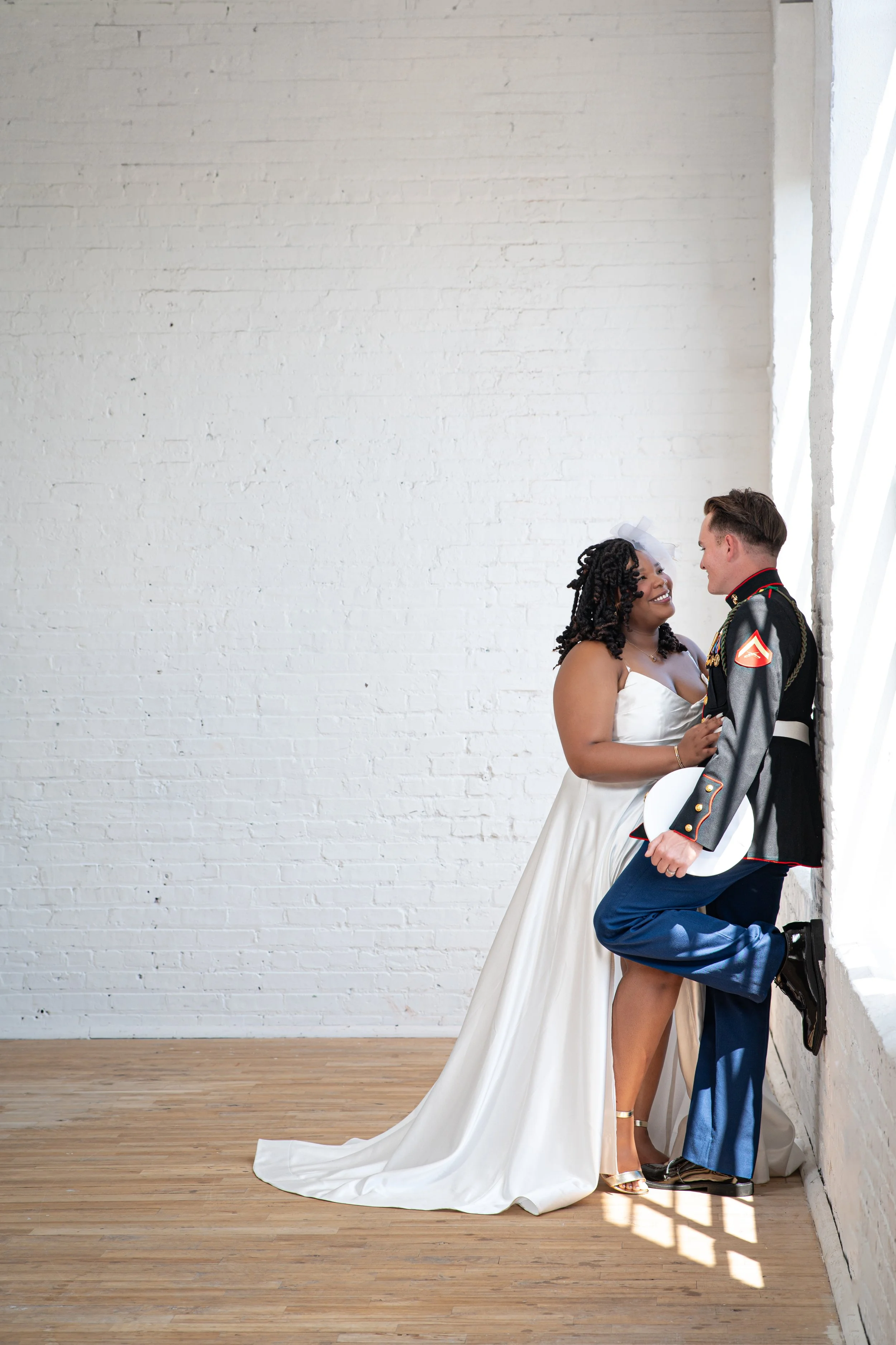 Bride leaning into her Marine groom in dress blues against a white brick studio wall